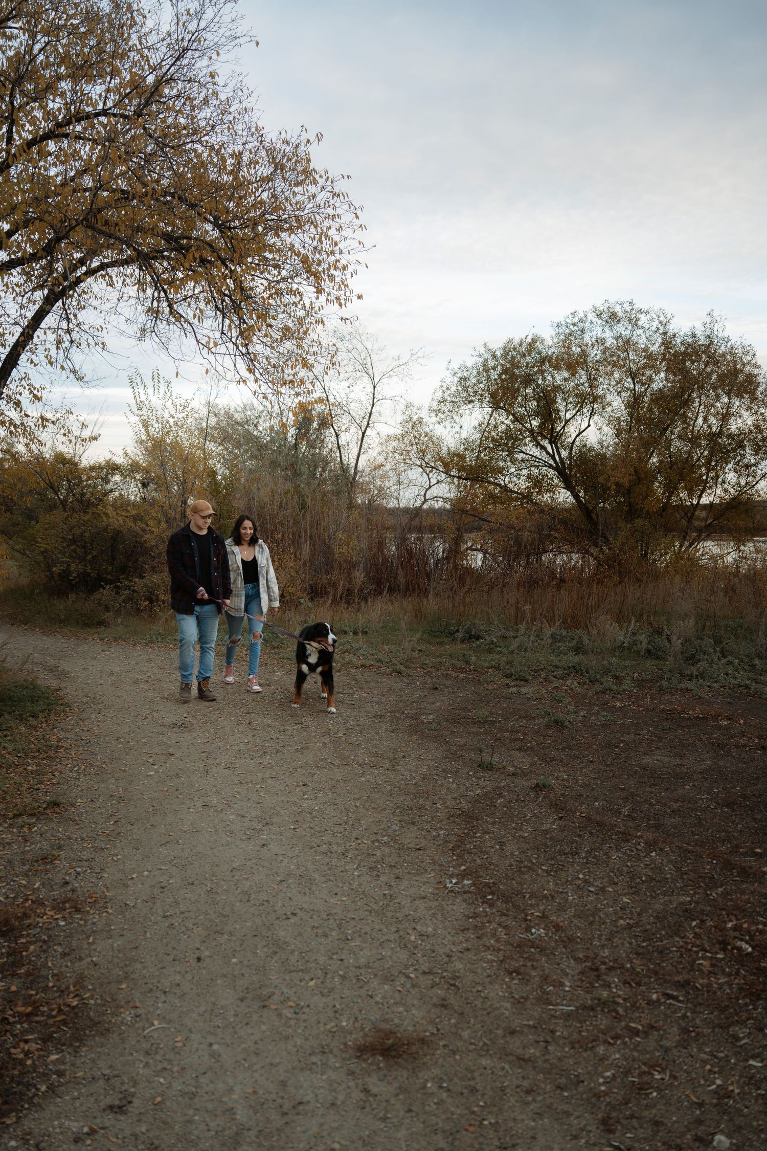 Couple walking a black and white dog on a dirt path surrounded by autumn trees near a lake.
