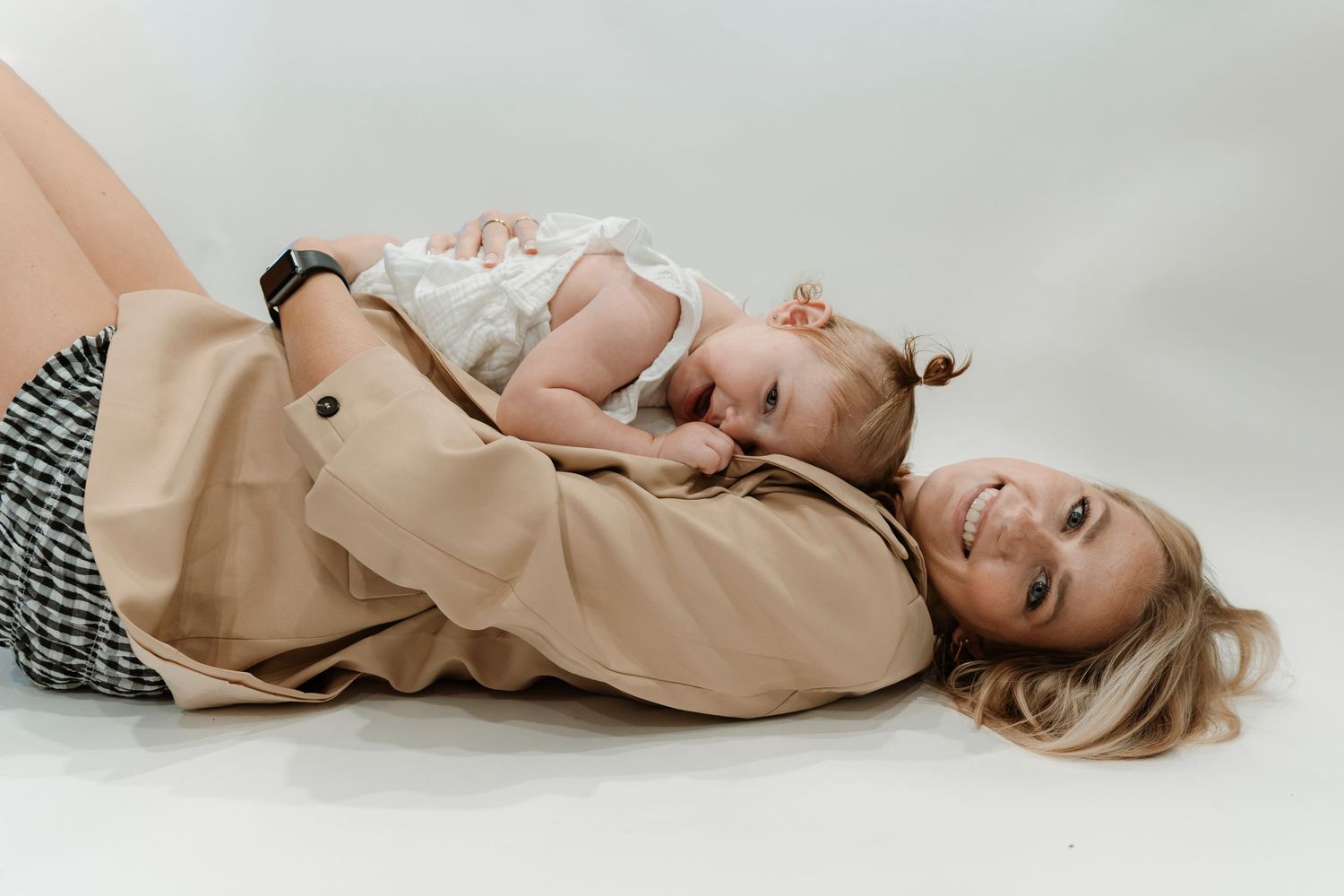 Mother lying on floor smiling while holding her baby girl in a white dress during a studio photo session.