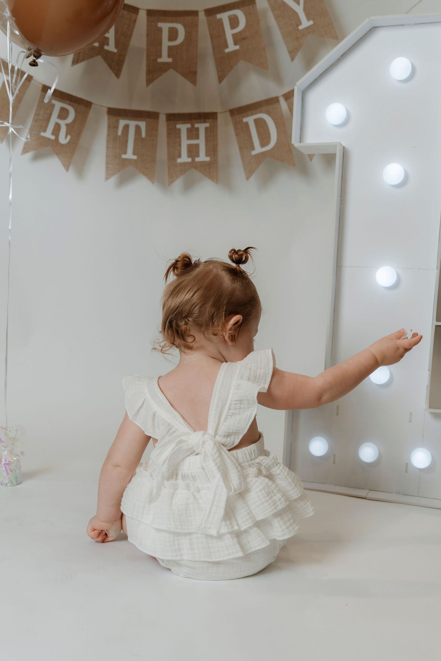 Baby girl in white dress sits before illuminated number 1 sign at first birthday party with Happy Birthday banner.