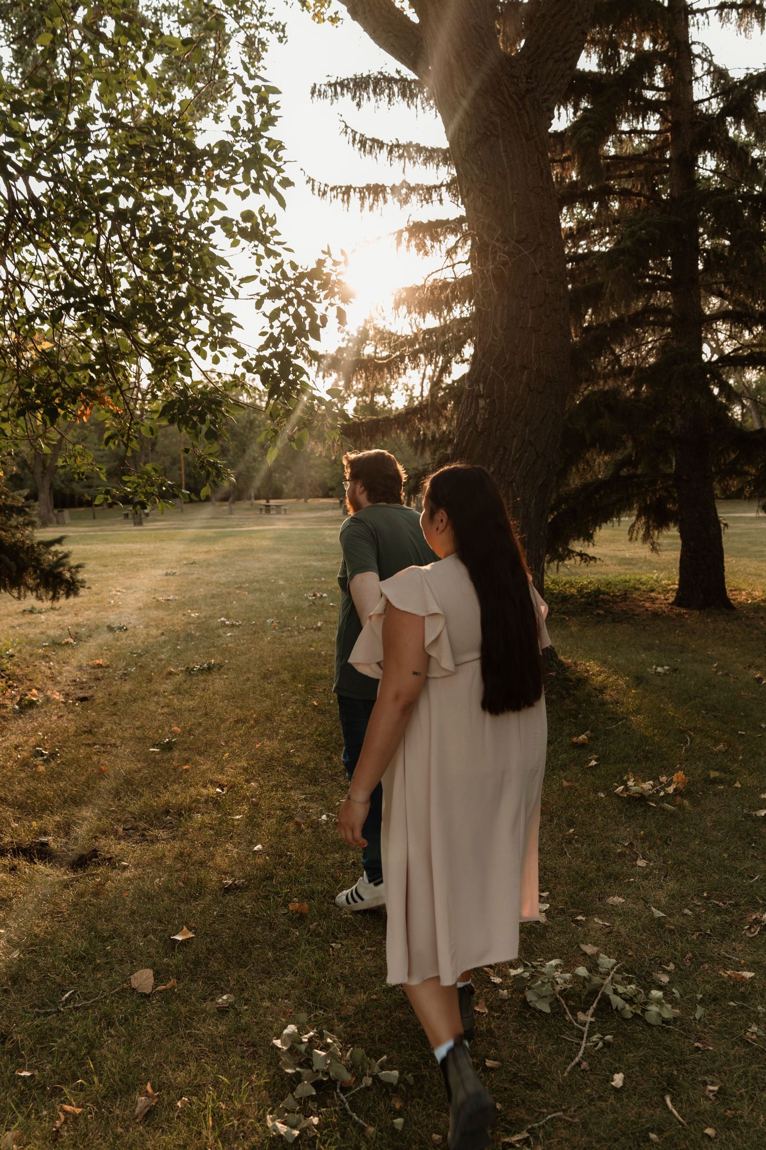 Couple walking hand in hand through a sunlit park with large trees during golden hour.