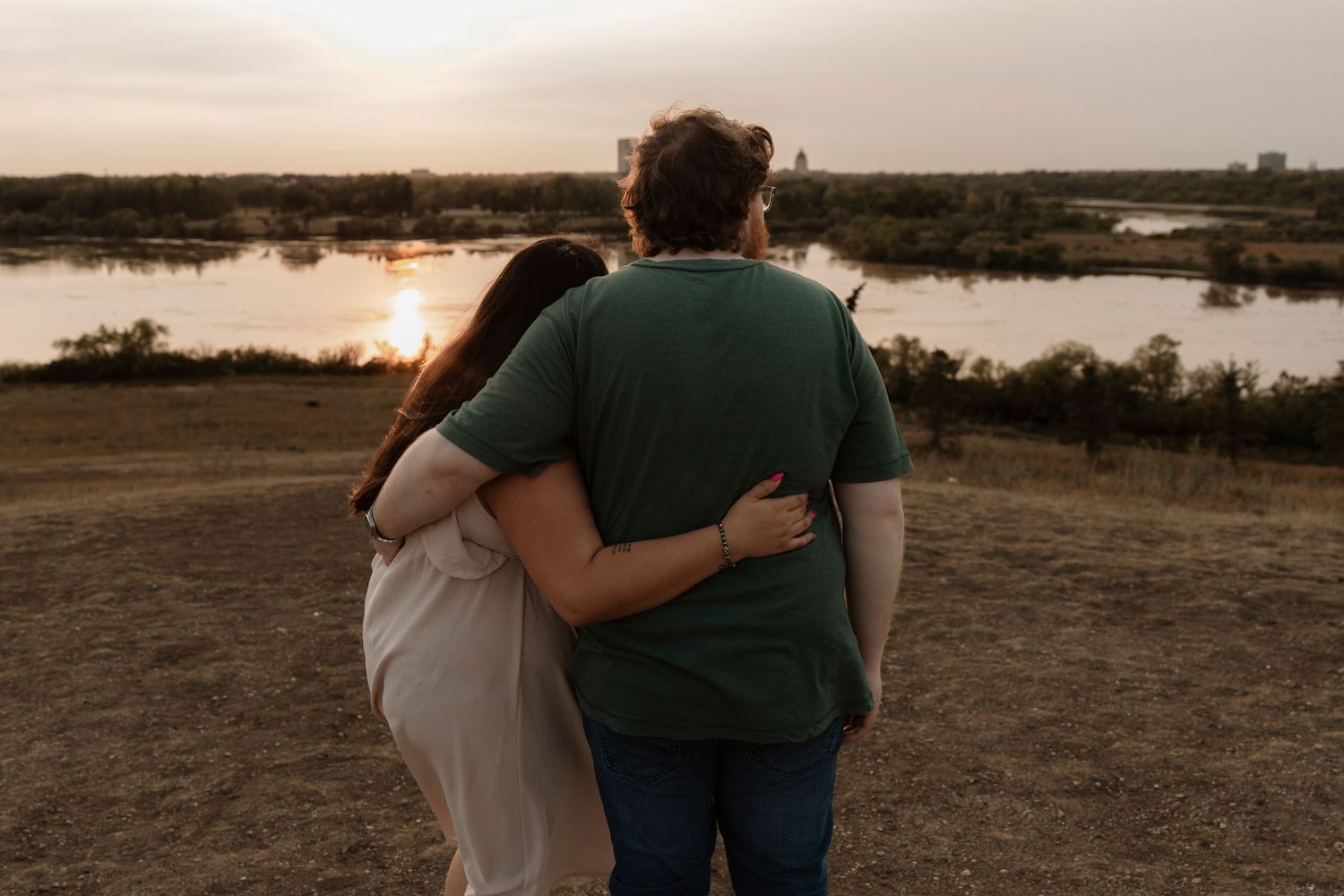 Couple embracing while watching a golden sunset over a serene river landscape in an open field.