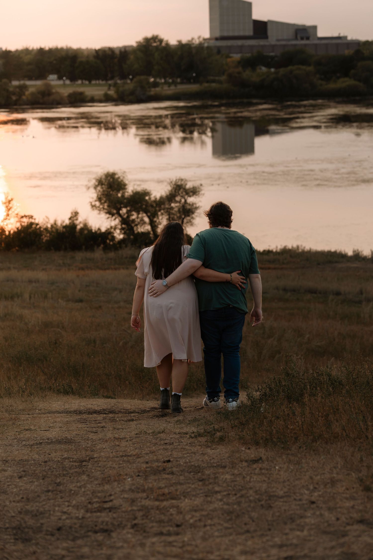 Couple walking arm in arm toward a serene lake at sunset, woman in white dress and man in green shirt.