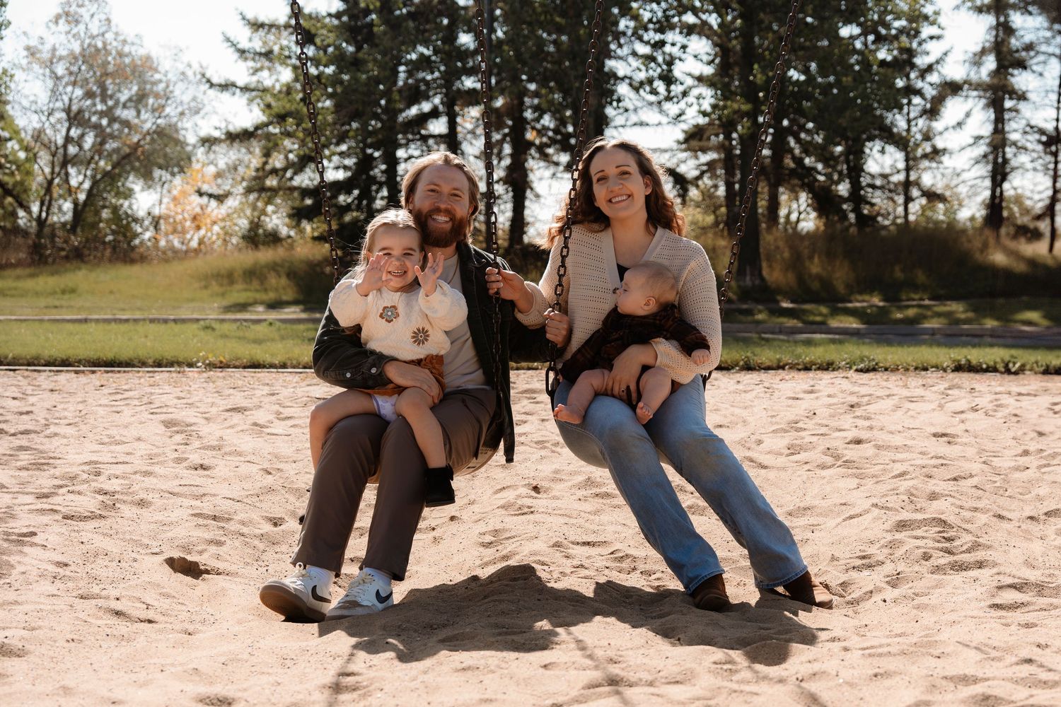Family of four with two young children sitting on sandy beach, smiling outdoors with trees in background.