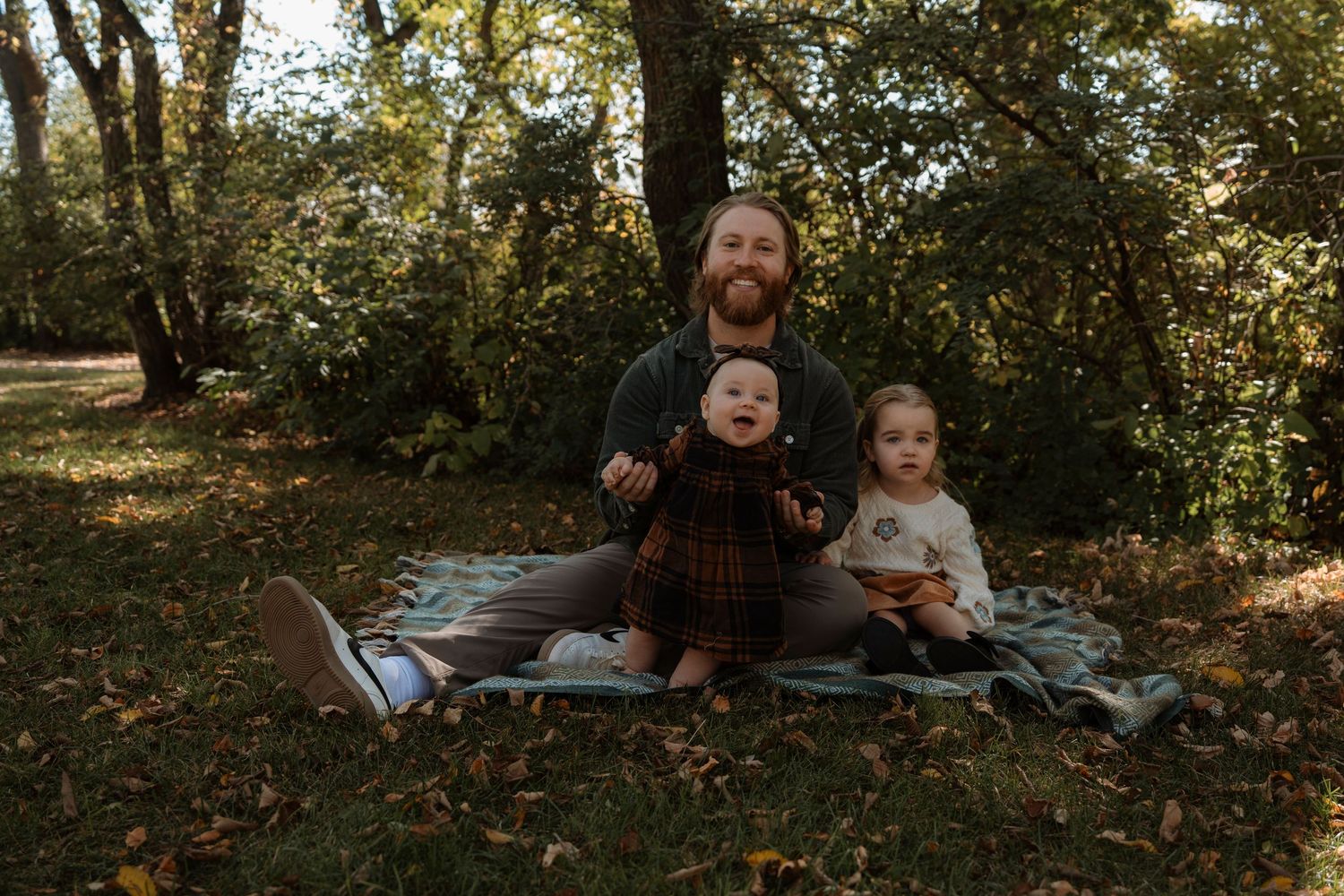 A bearded father sits on a blanket outdoors with two young toddlers among autumn trees and fallen leaves.