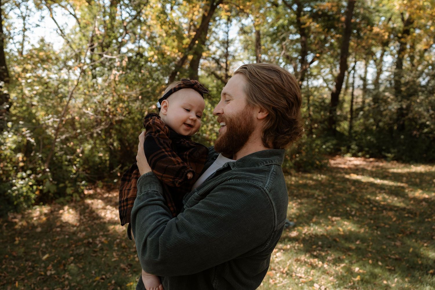 A bearded man holds a smiling baby outdoors in an autumn forest setting with golden foliage.