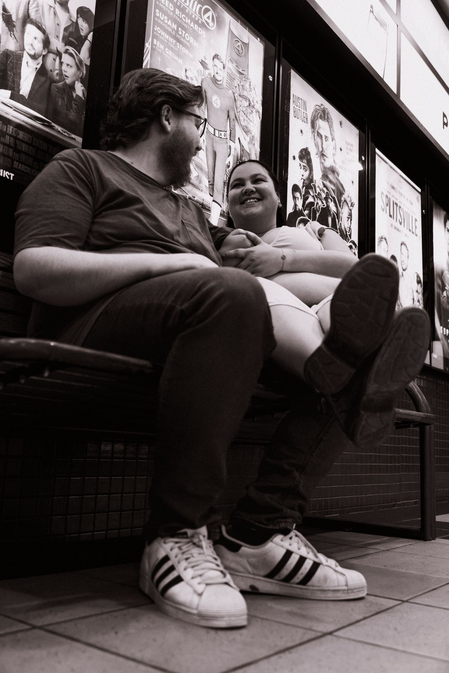 Black and white photo of a couple sitting and laughing together outside a theater with movie posters.