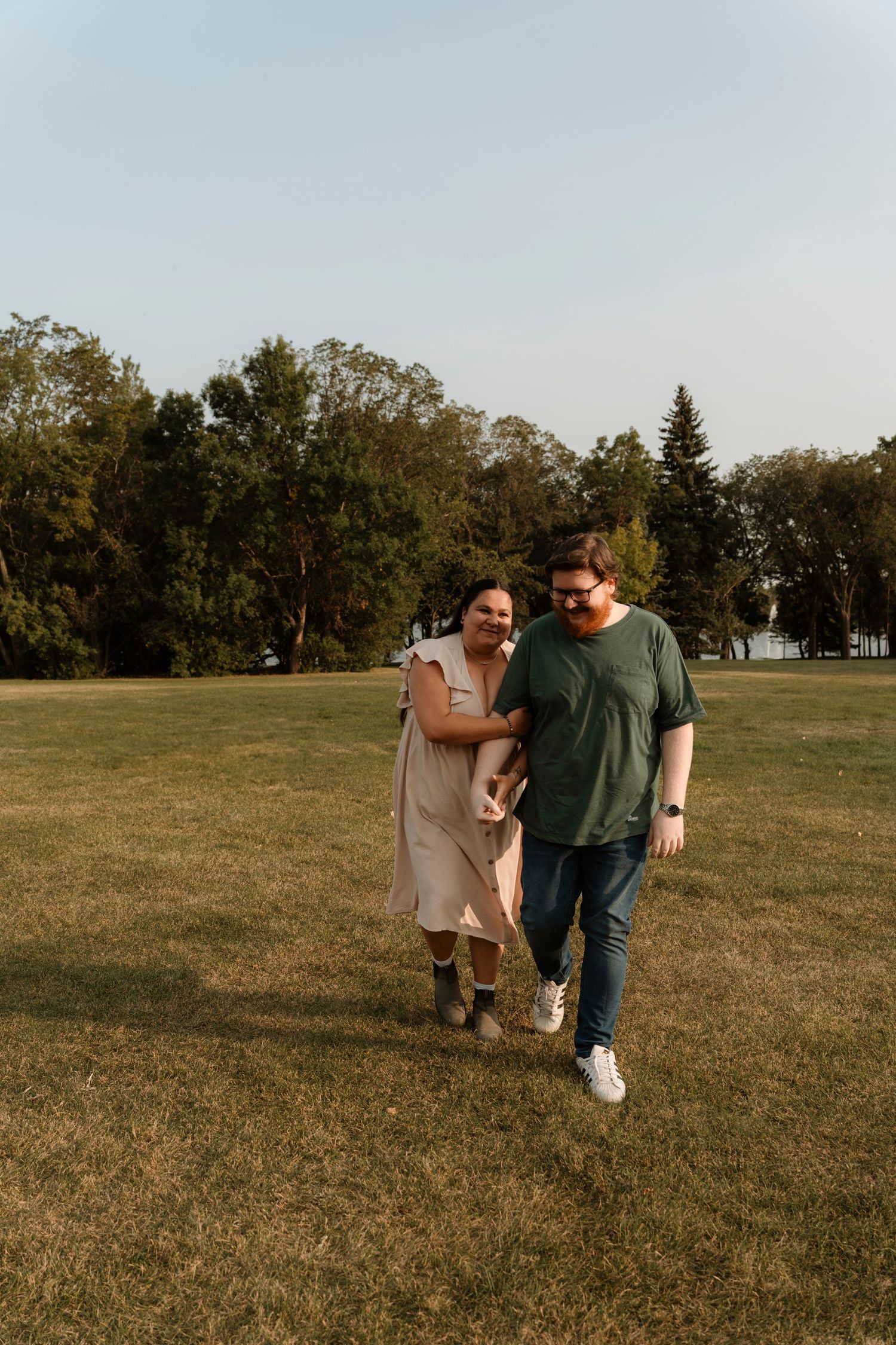 Couple walking arm in arm through a green park, smiling, wearing casual summer outfits with trees in the background.