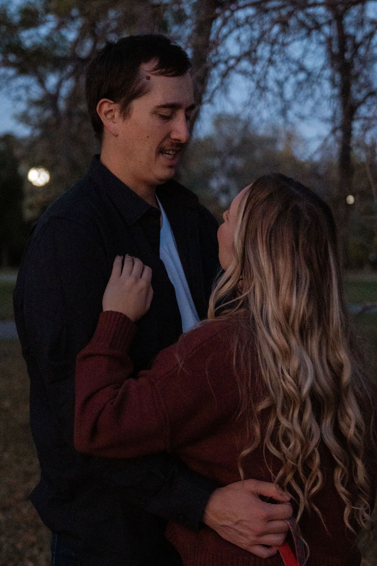 Couple sharing a romantic moment at dusk in a park, woman in burgundy sweater embracing man in dark jacket.