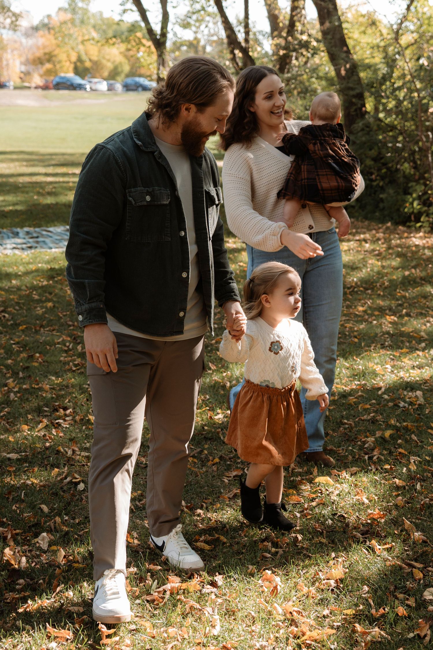 A young family with two children enjoys an autumn walk in a park, surrounded by fallen leaves and trees.