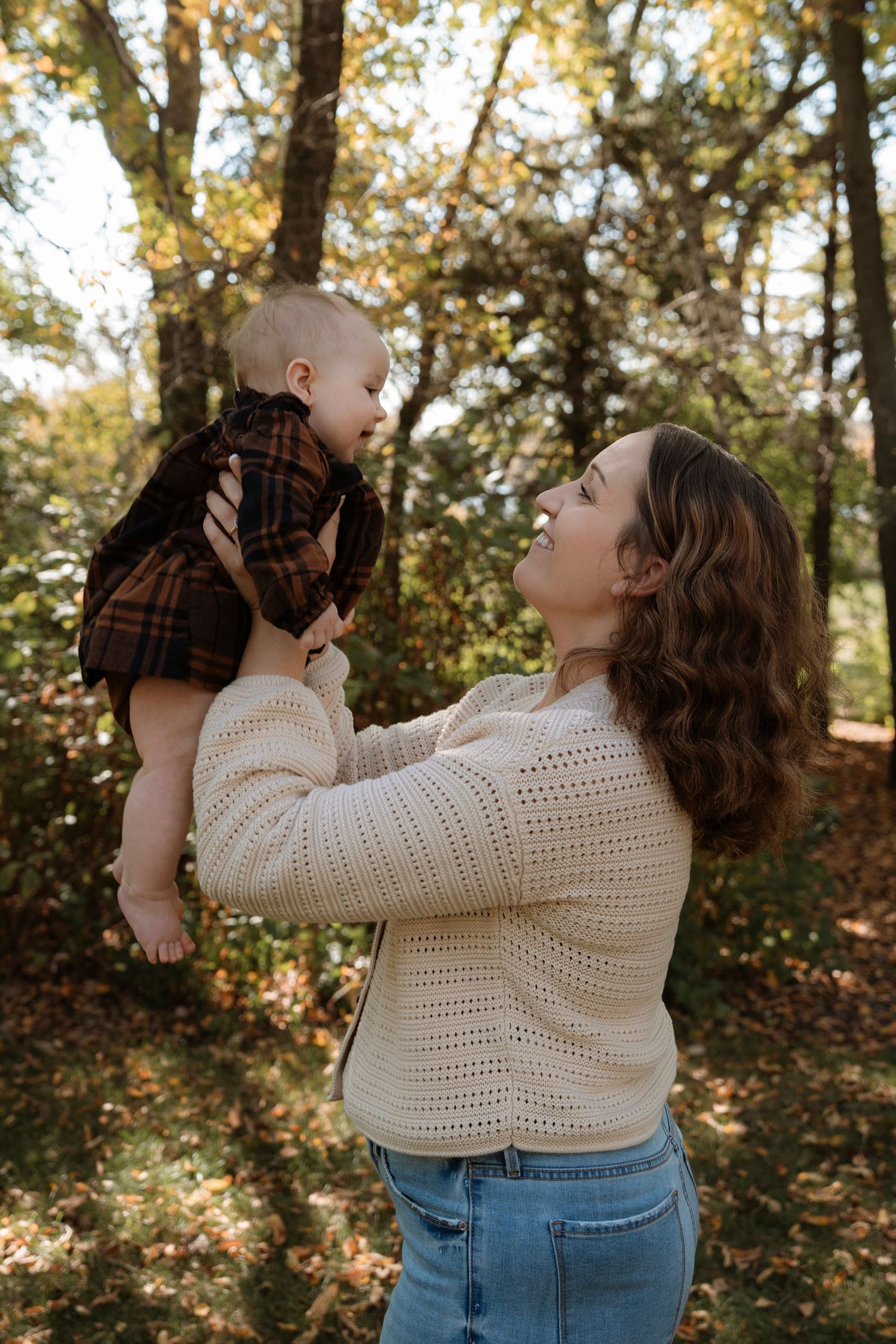 A smiling young woman in a cream sweater lifts a baby in a plaid outfit outdoors in an autumn forest setting.