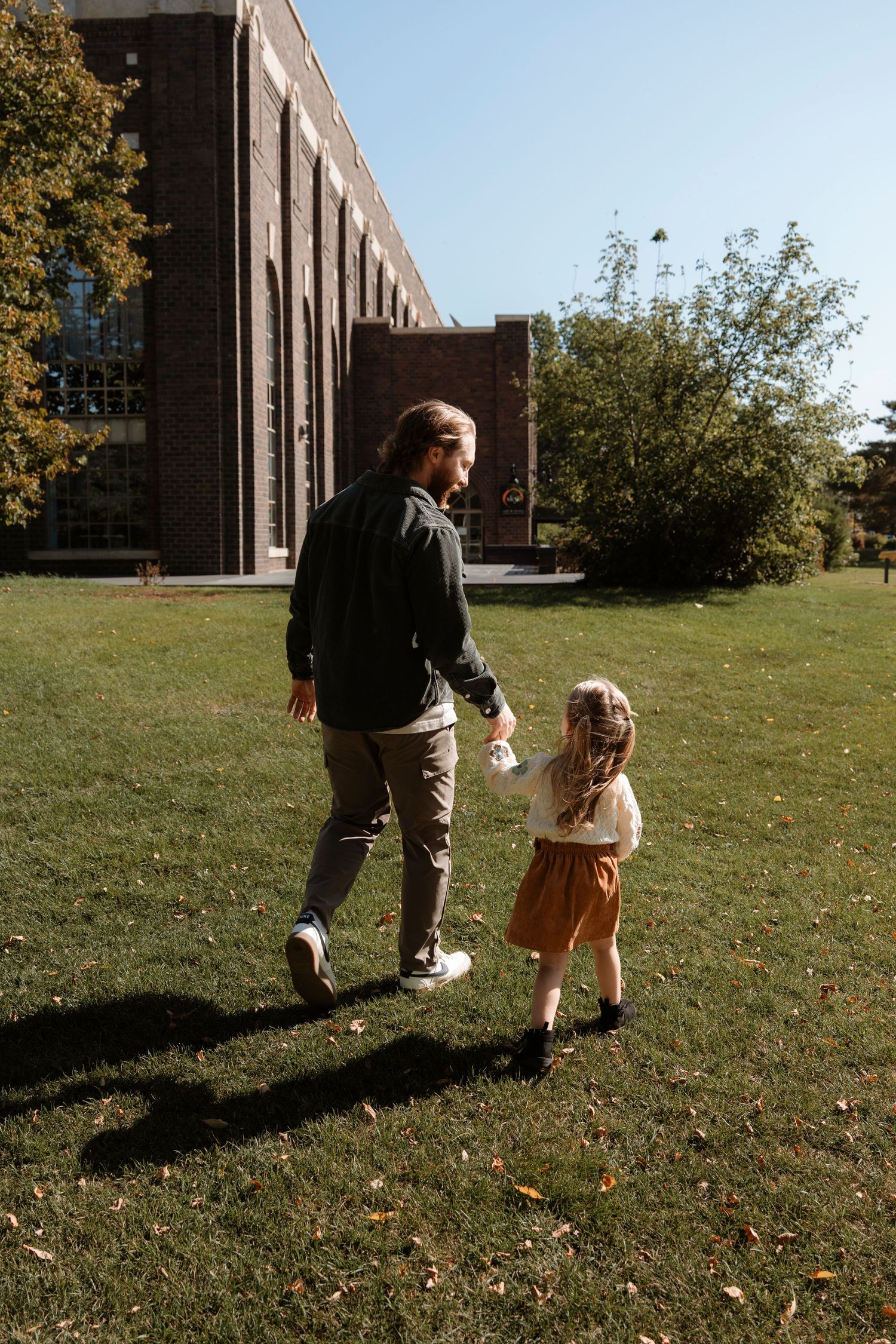 A father and young daughter walk hand-in-hand across a sunny green lawn near a brick building.