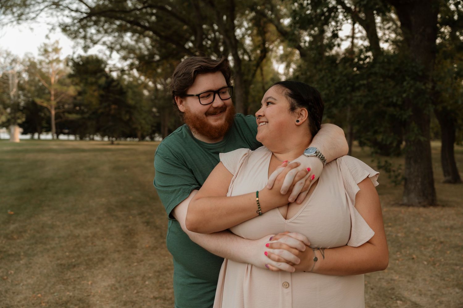 Couple embracing and laughing together in a lush green park during an outdoor engagement photo session.