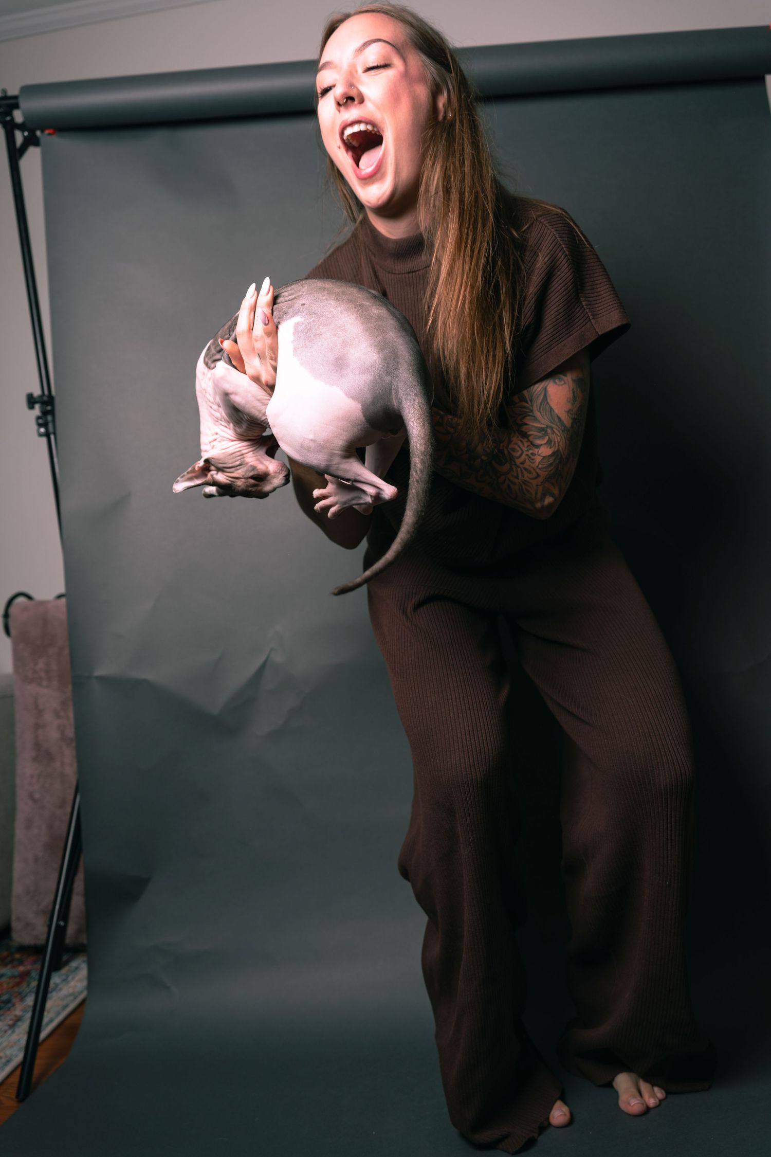 Woman in brown outfit holding a hairless Sphynx cat while laughing in a photography studio setting.