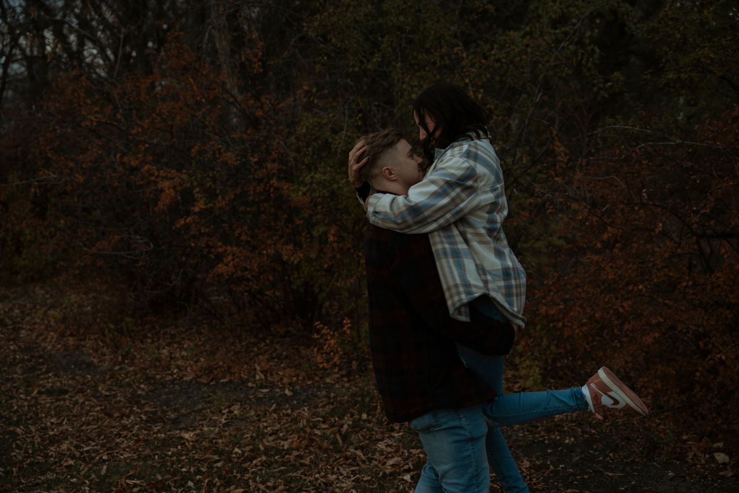 A couple embraces romantically in an autumn forest, man lifting woman among colorful fallen leaves and dark trees.