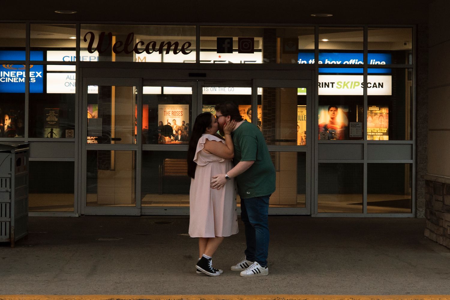 Couple sharing a romantic kiss outside a lit welcome sign storefront at dusk.