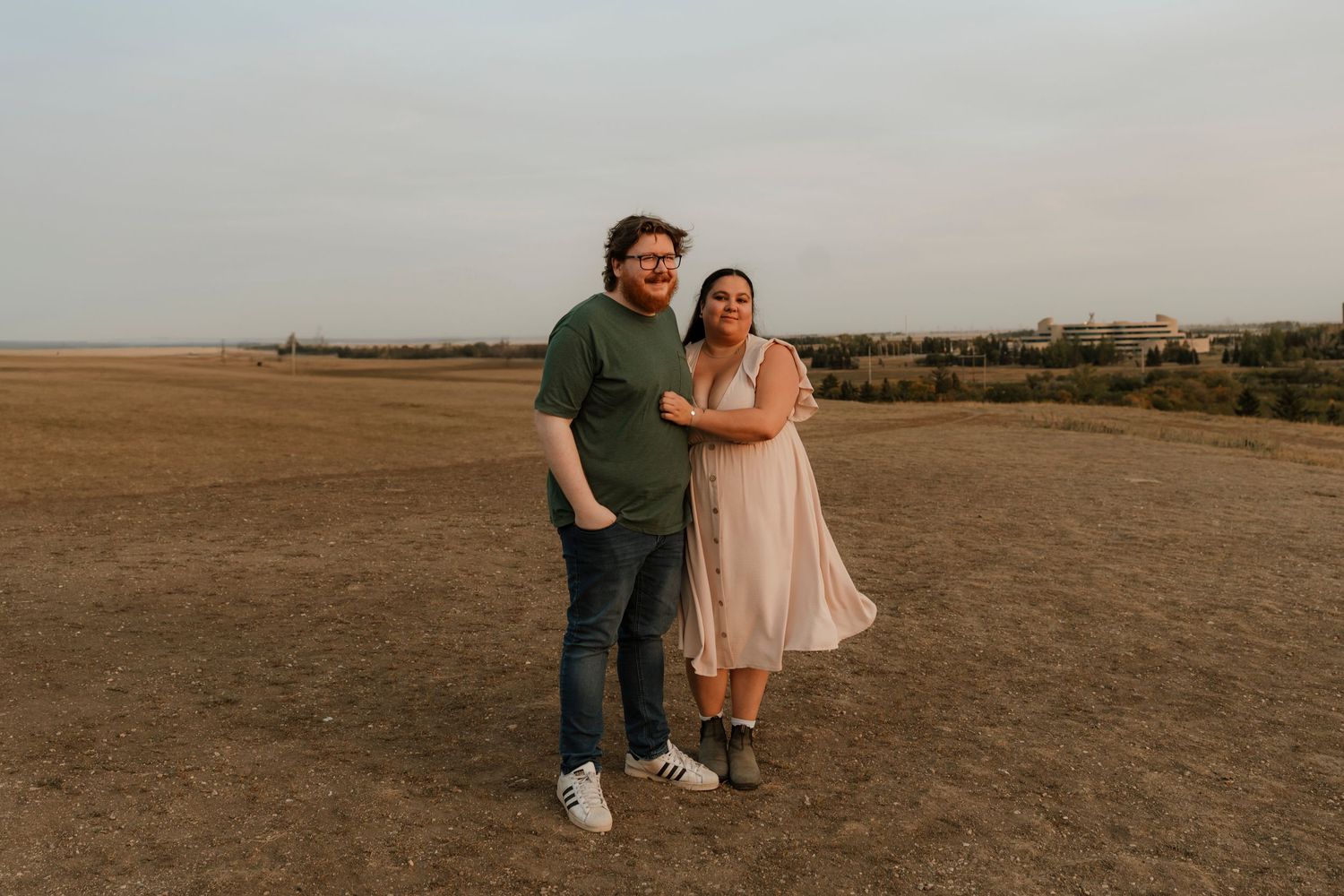 Couple posing together on open field at golden hour, man in green shirt and woman in pink dress smiling outdoors.