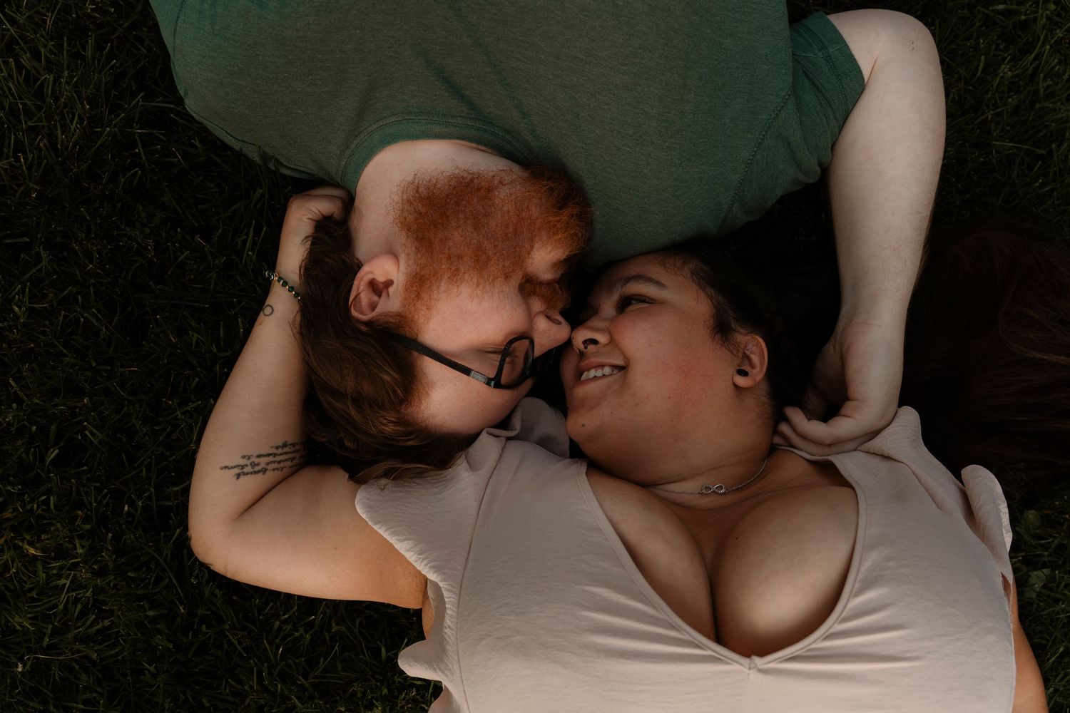 Two people lying on grass, facing each other and smiling intimately, one with red hair wearing glasses.