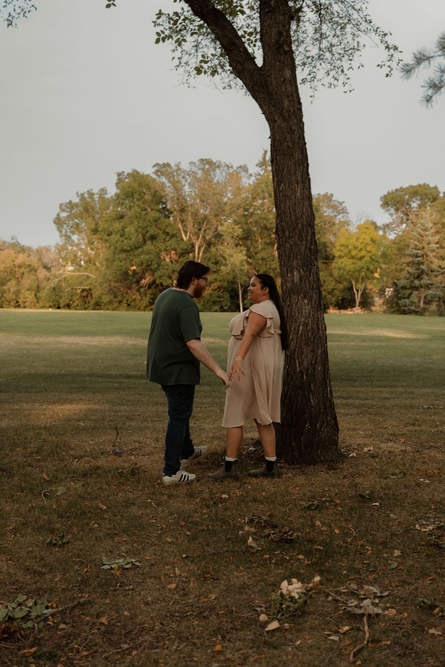 A couple holds hands near a large tree in a lush green park, sharing a romantic moment outdoors.