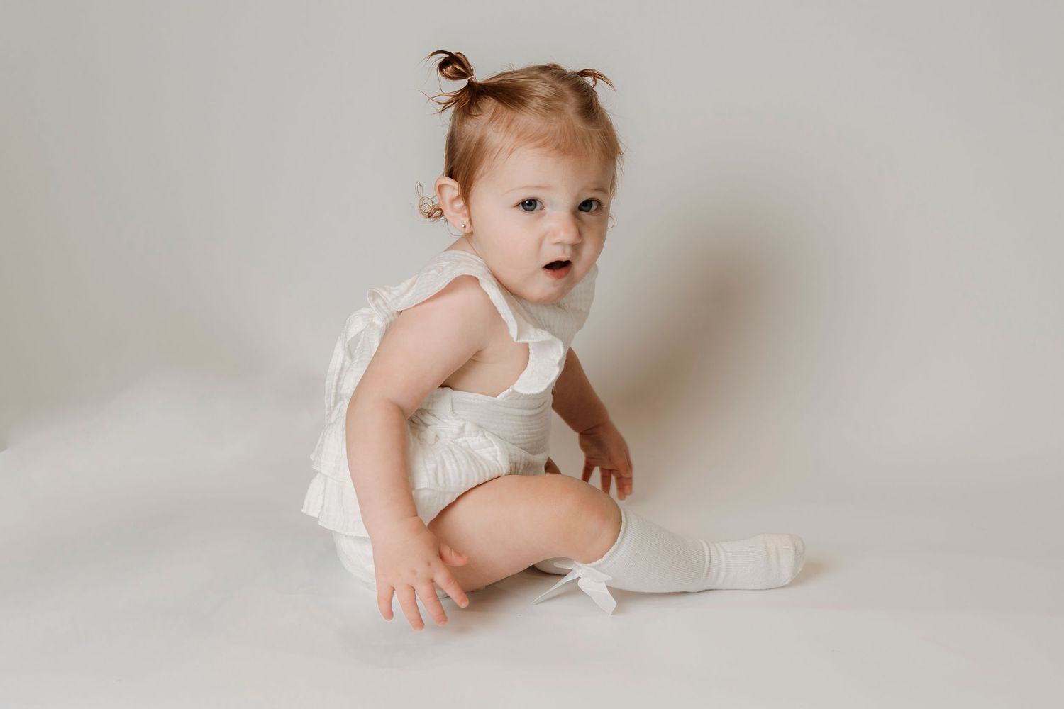 A cute baby girl in a white romper and knee socks sits on a white background, looking curiously to the side.