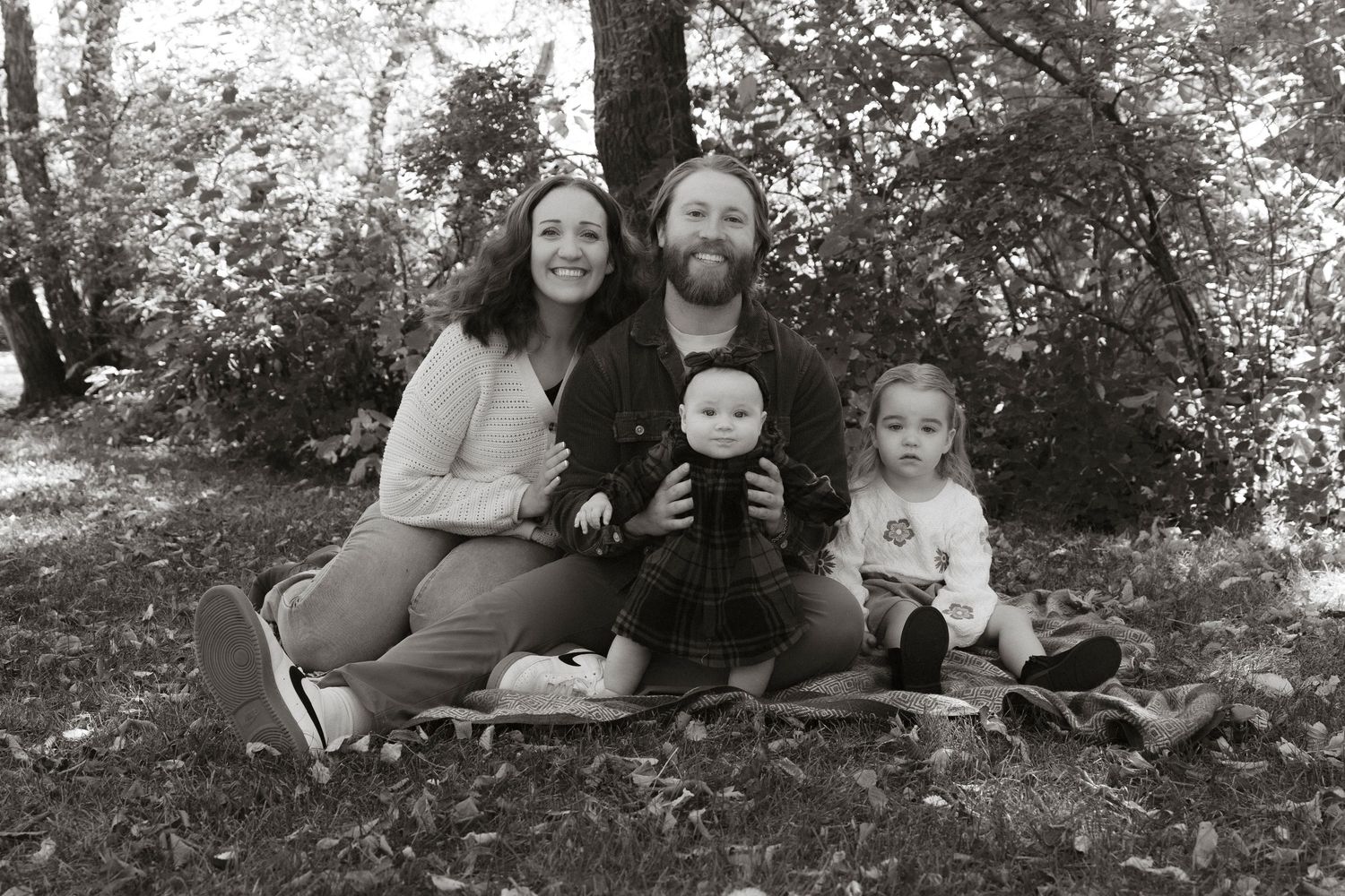 Black and white family portrait of parents with baby and toddler sitting outdoors on blanket among trees.