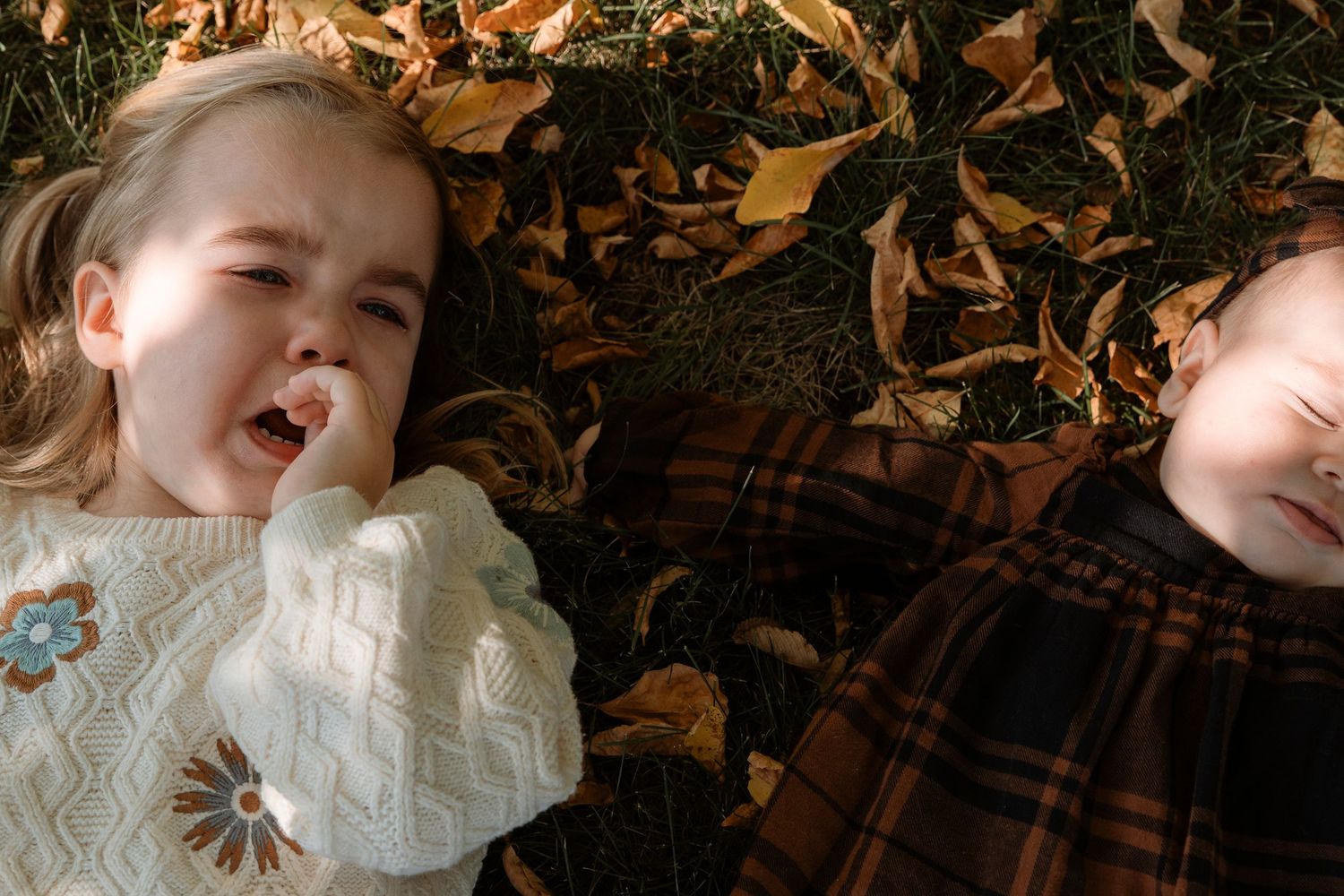 Two young children lying on grass covered with autumn leaves, one laughing and one resting eyes closed.