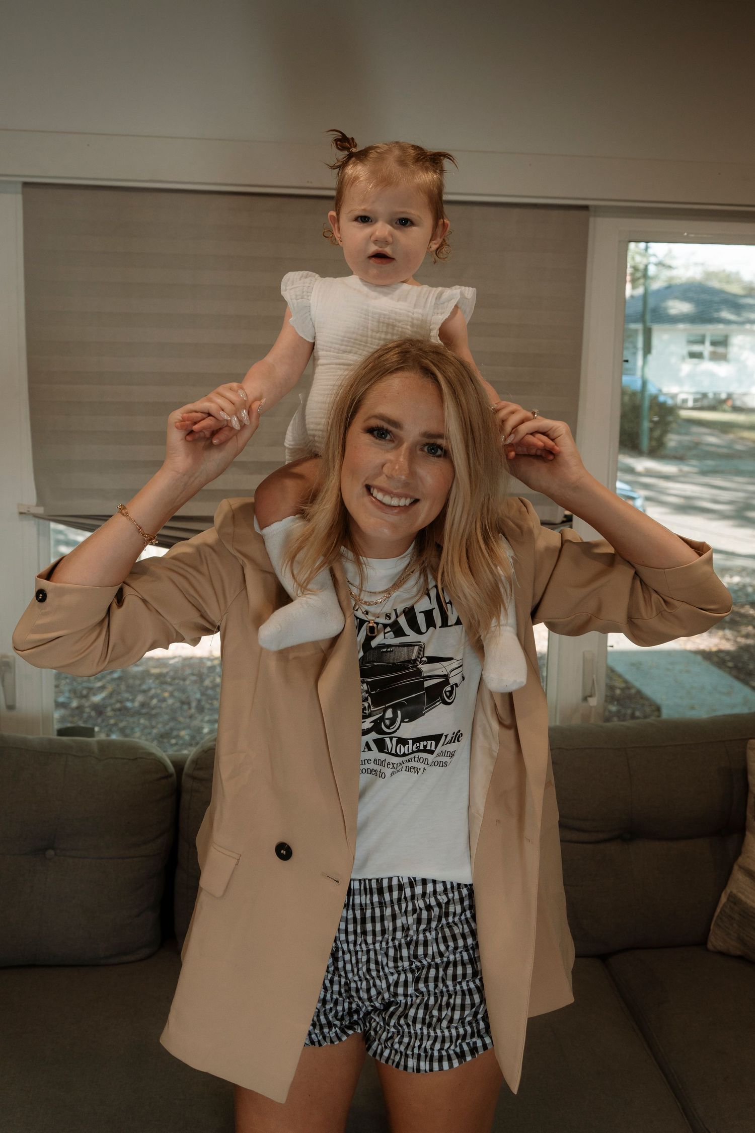 A smiling blonde woman carries a baby girl on her shoulders in a cozy living room, wearing a tan blazer and graphic tee.