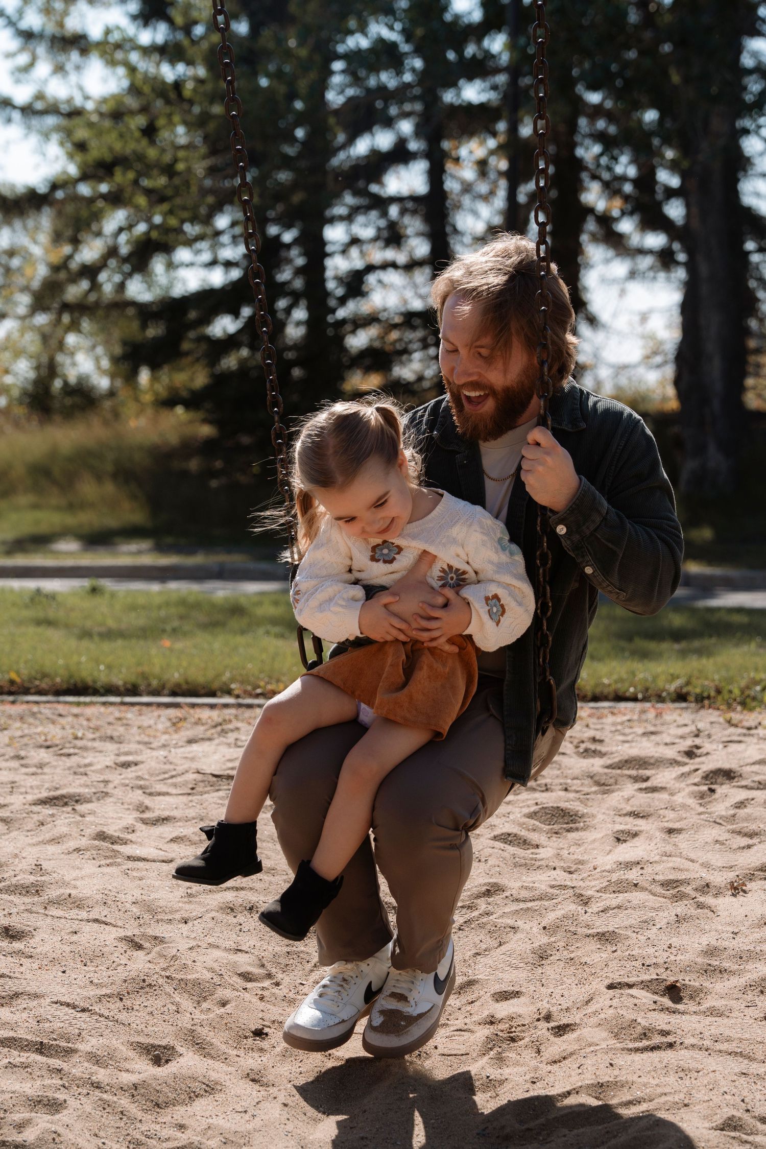Bearded father sitting on playground swing holding young daughter in sandy park with trees in background.