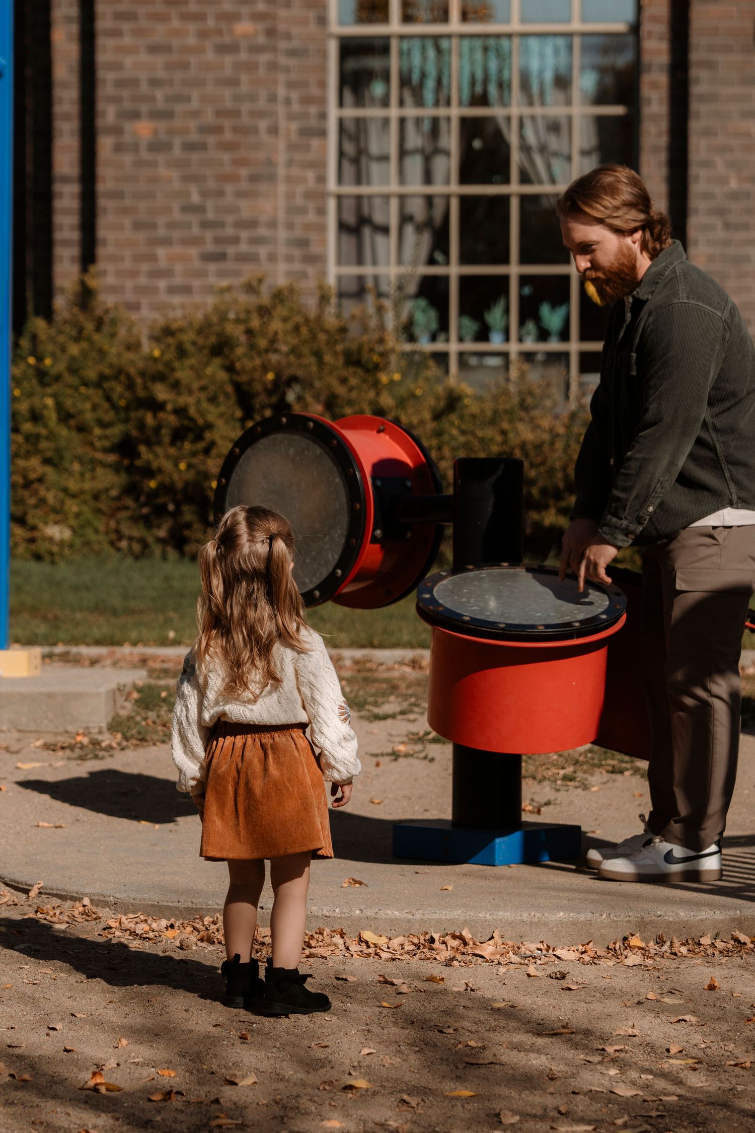 A man and young girl explore a red outdoor musical drum instrument on a sunny day near a brick building.