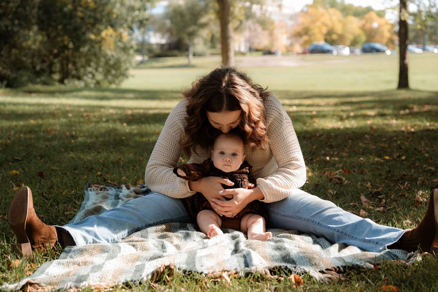 A mother sits on a blanket in a park, lovingly hugging her baby in autumn sunlight.