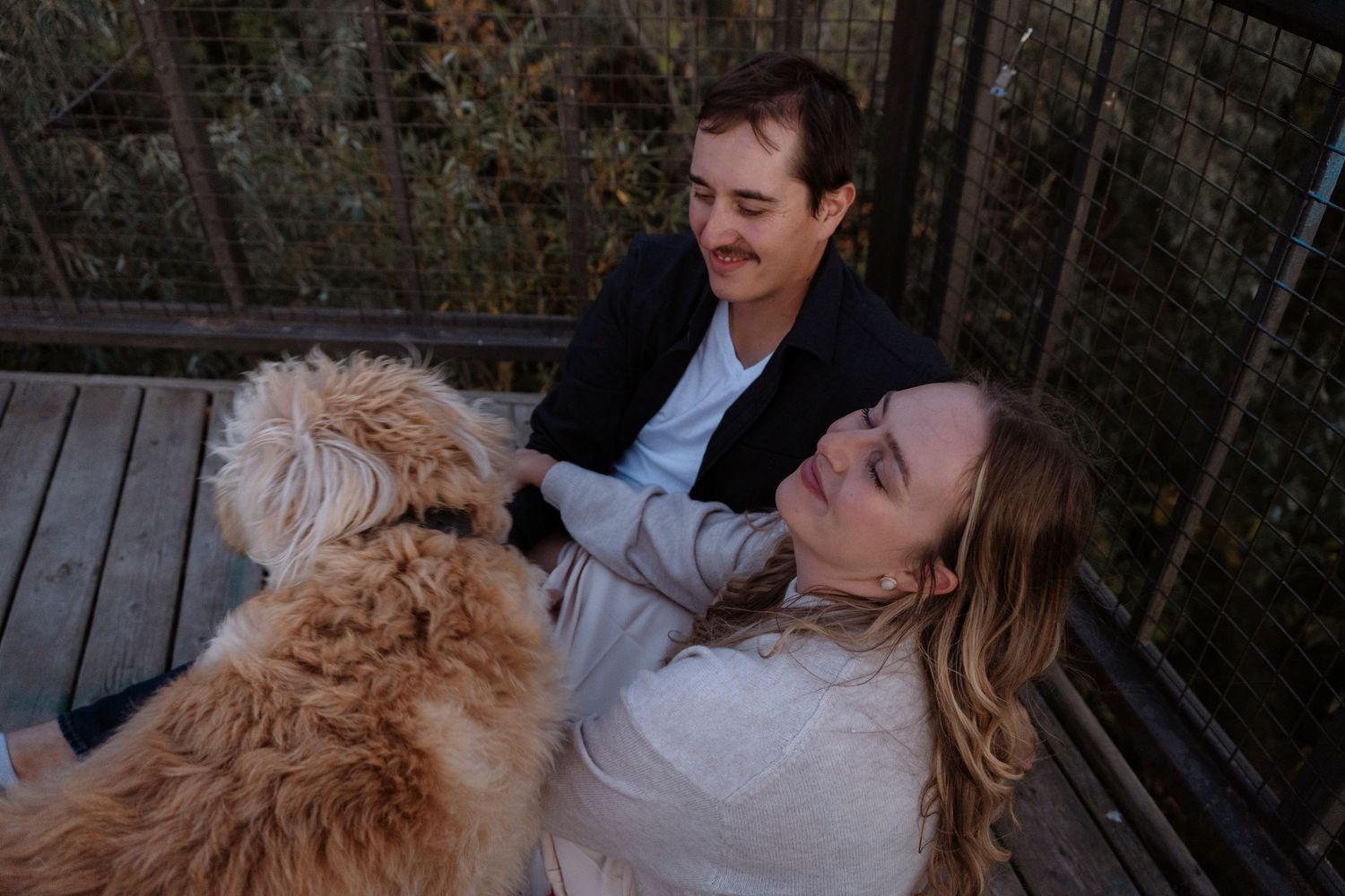 Two people and a fluffy golden dog relaxing together on a wooden deck surrounded by nature.