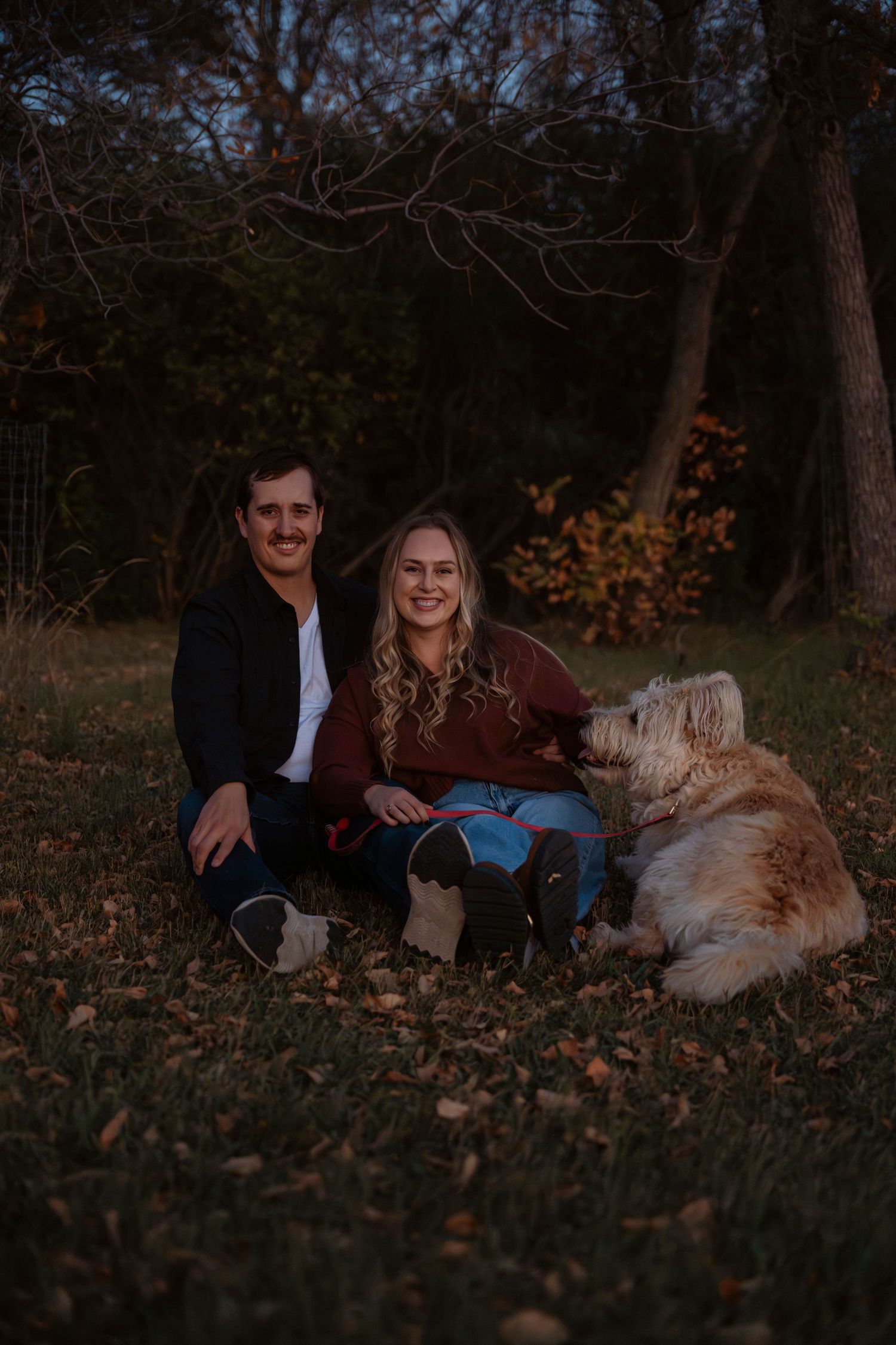 A smiling couple sits on grass with a fluffy golden dog beside them, surrounded by autumn trees at dusk.