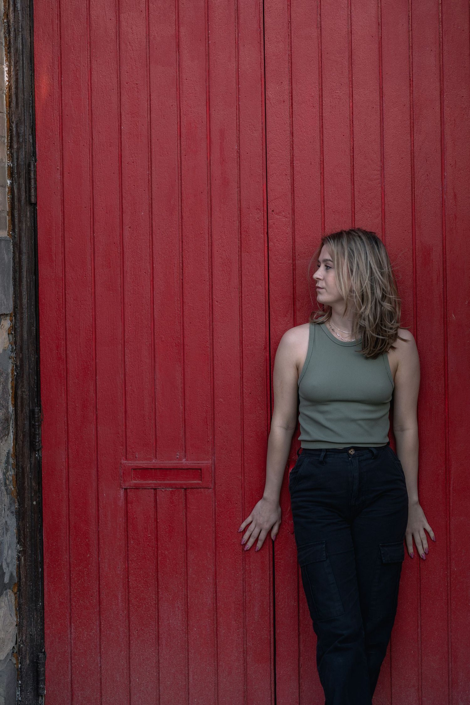 Young woman in green tank top and black jeans leaning against a bold red wooden door outdoors.