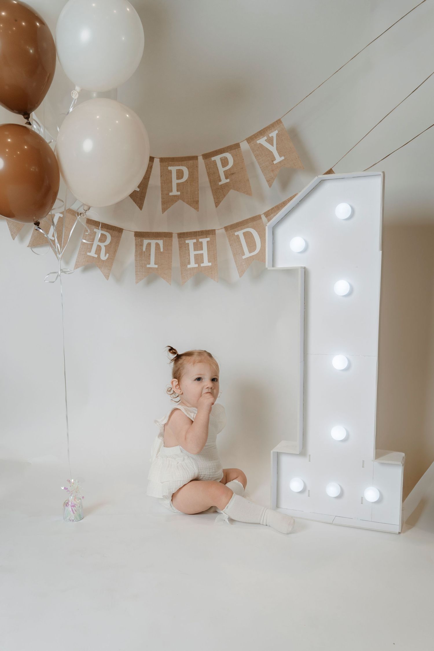 Baby girl sitting beside illuminated number 1 sign with Happy Birthday bunting and brown and white balloons.