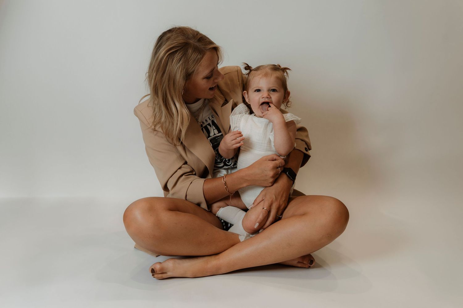 A smiling blonde woman sits cross-legged on a white backdrop, lovingly holding a happy baby girl in a white outfit.