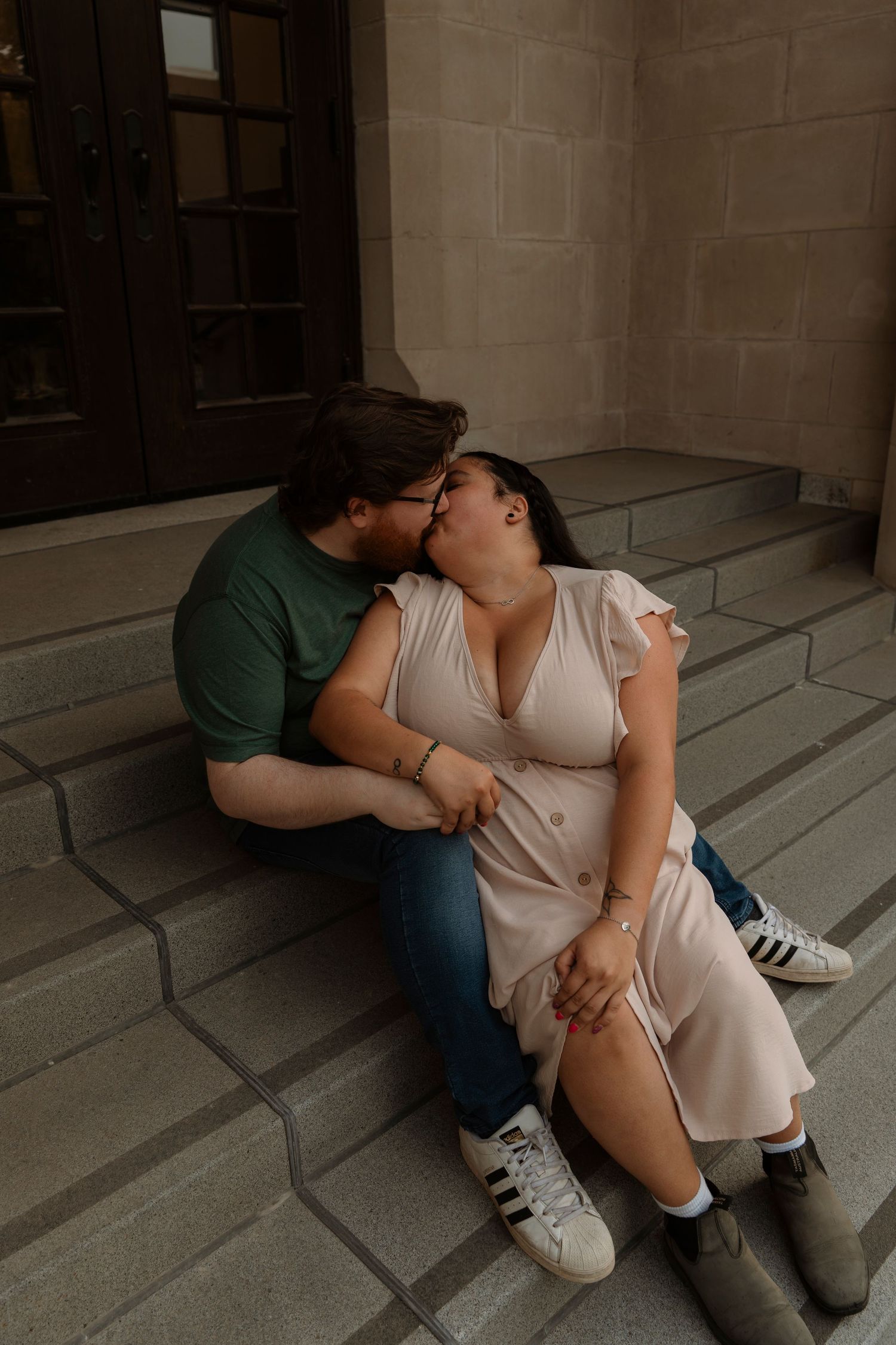 Couple sharing a romantic kiss while sitting on stone steps outside a building, wearing casual outfits.