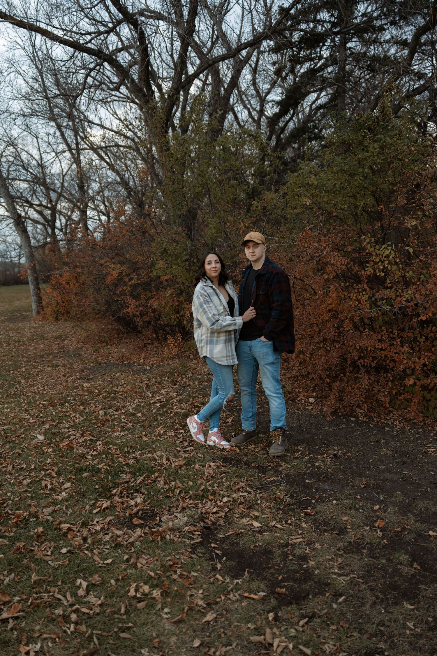 Couple standing together in autumn park with bare trees and orange foliage in background.