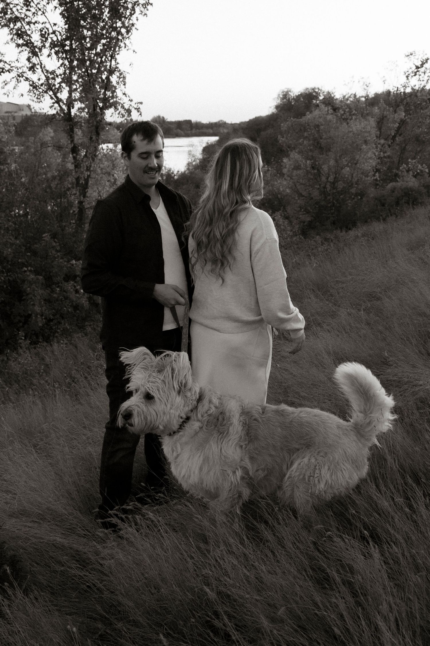 Black and white photo of a couple walking with a fluffy white dog through a scenic outdoor field near a lake.