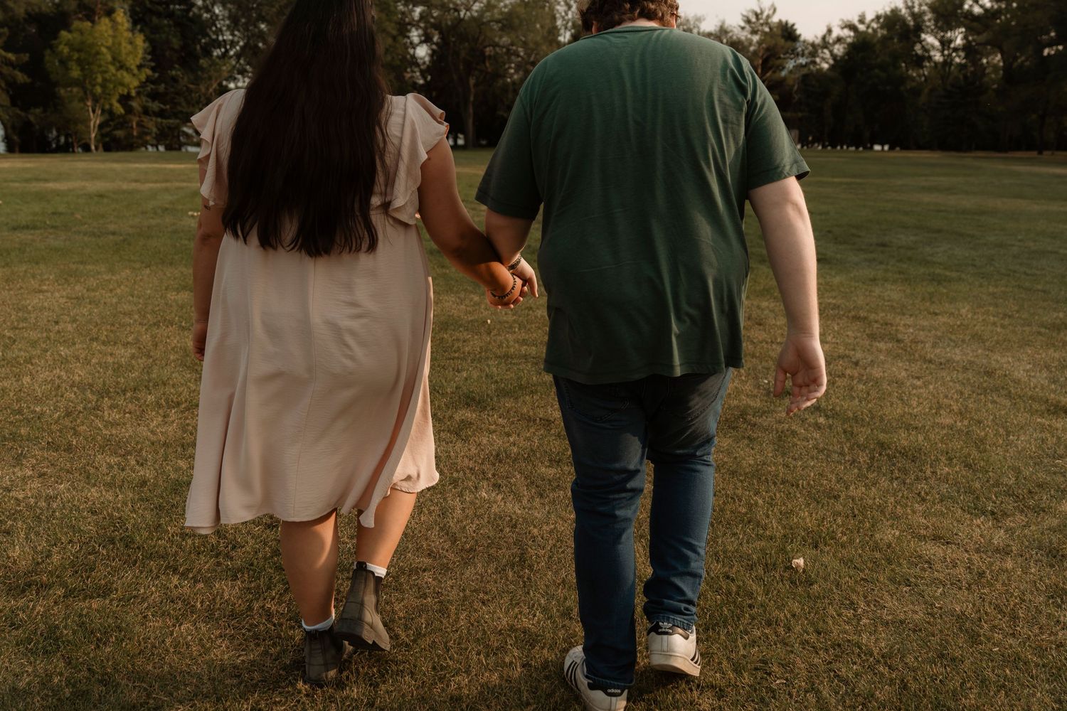 Couple holding hands walking through a green park, woman in white dress and man in green shirt, romantic outdoor stroll.
