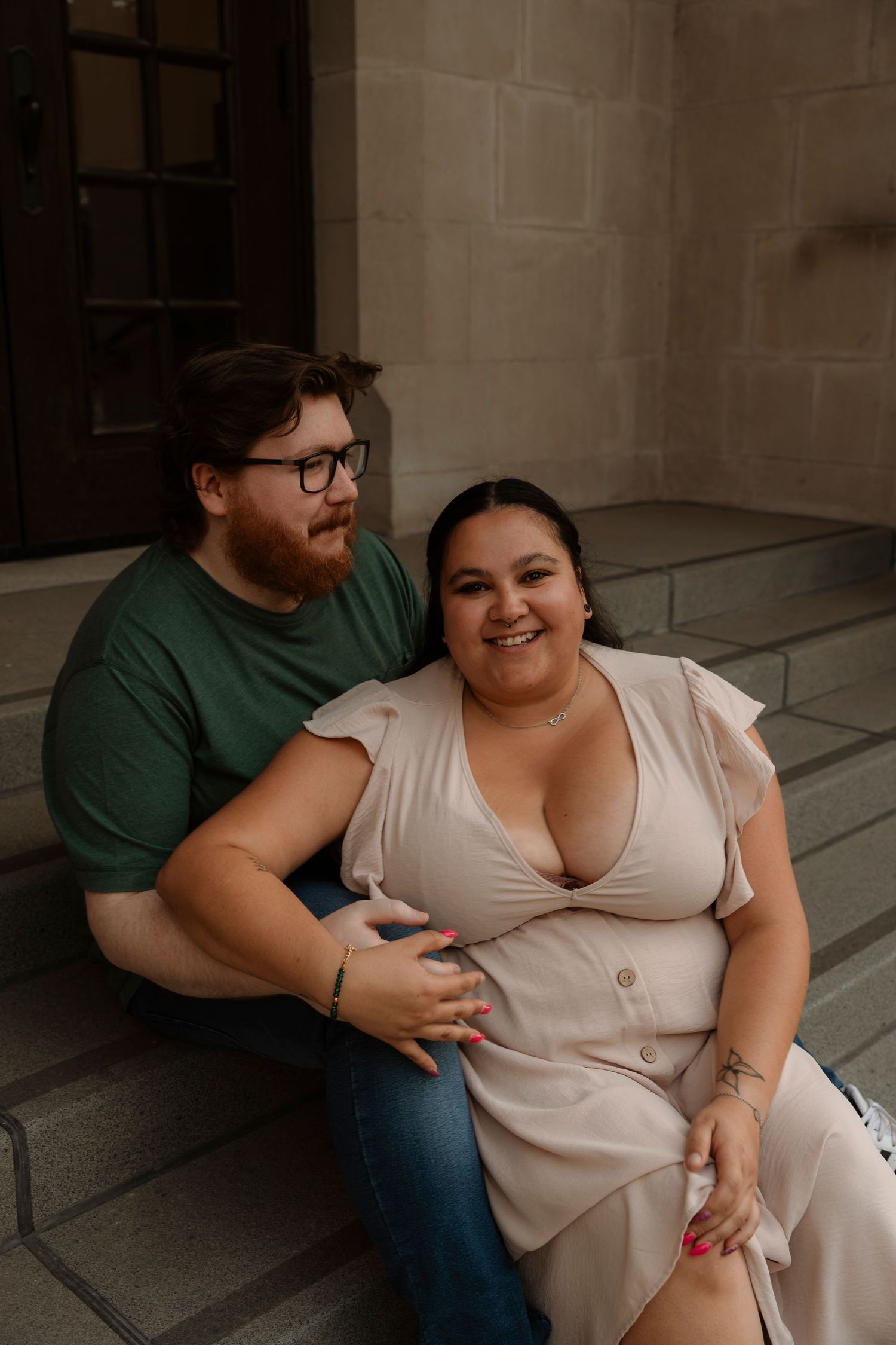 A couple sits together on stone steps, smiling, man in green shirt with glasses, woman in light pink dress.