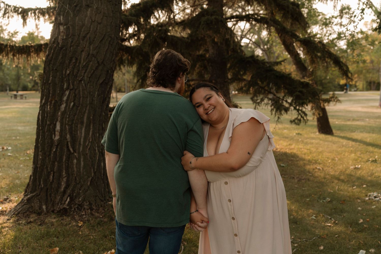 Couple embracing near a large tree in a park during golden hour, woman smiling warmly in cream dress.