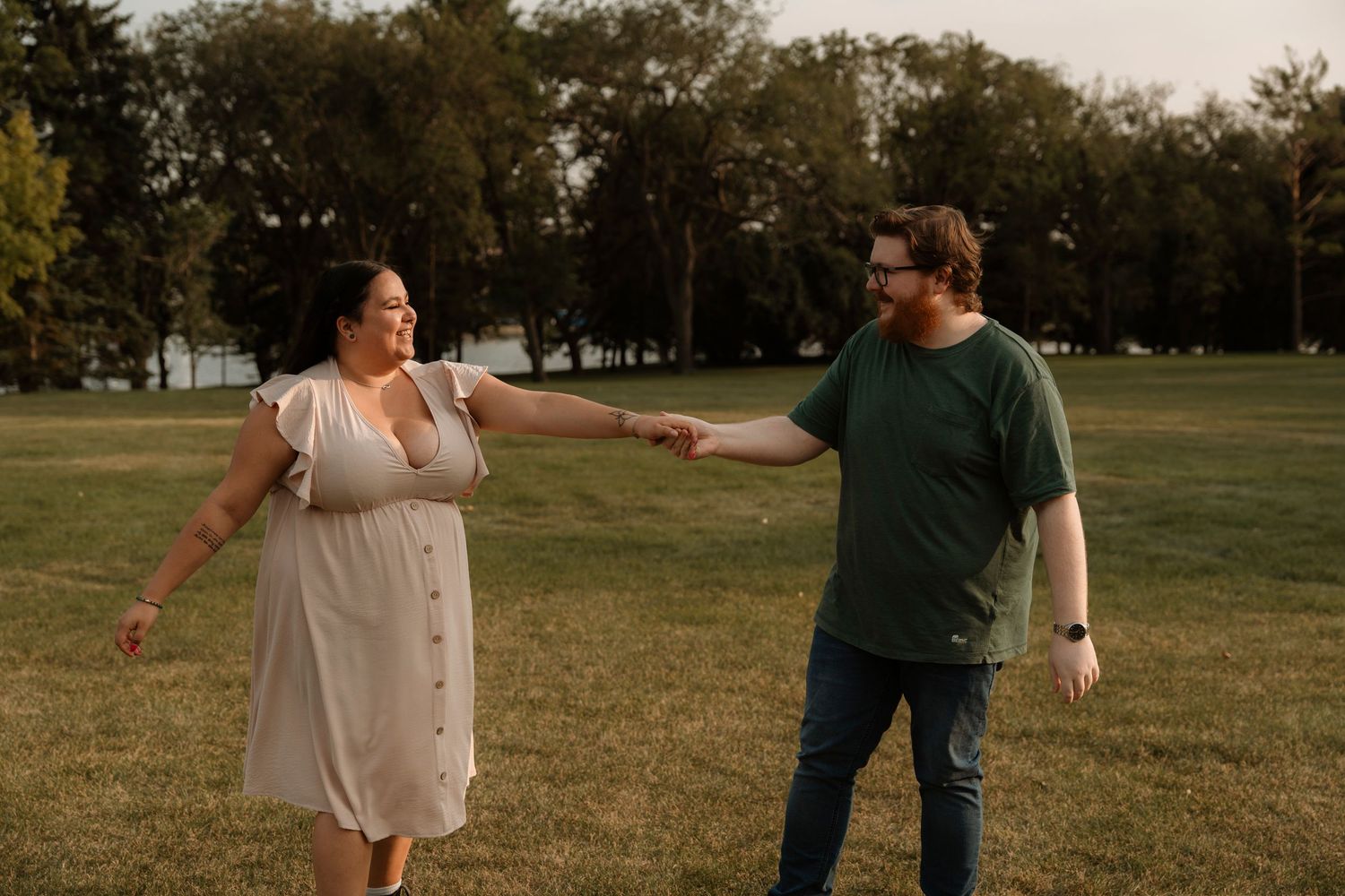 Couple holding hands in a green park, woman in white dress and man in green shirt, golden hour outdoor photo.