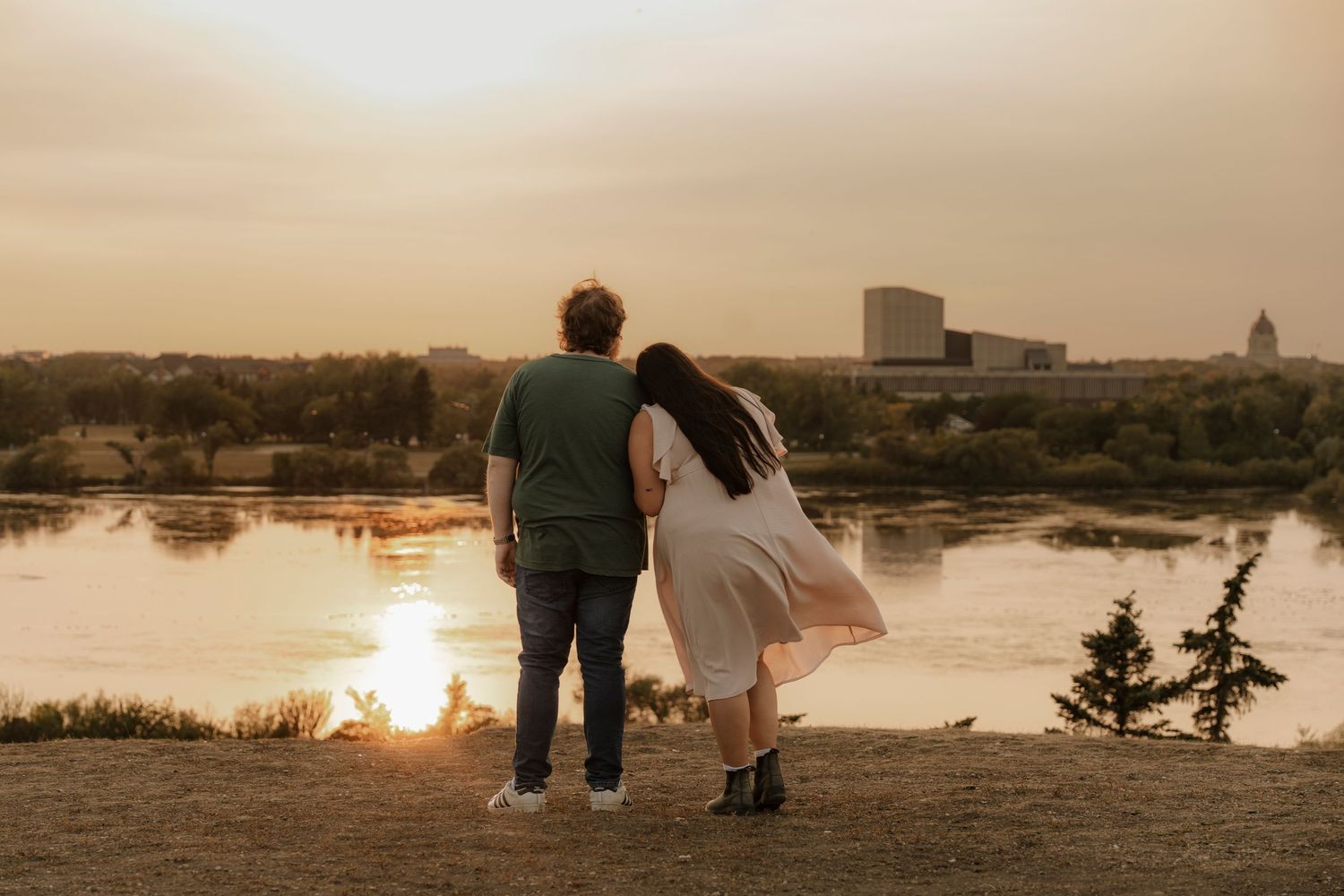Couple standing together watching a golden sunset over a calm river with a city skyline in the background.