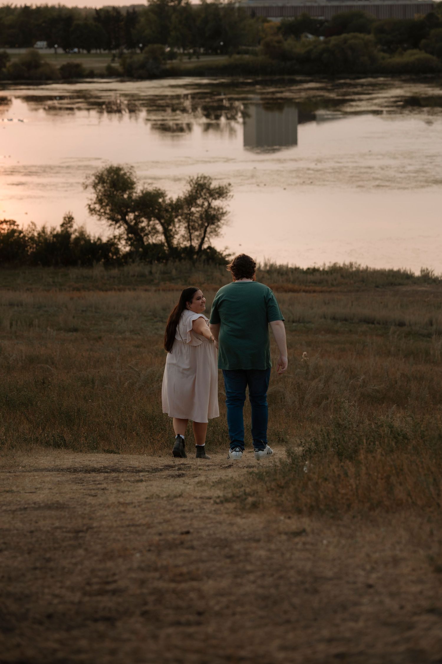 Couple walking hand in hand along a scenic riverside path at sunset, woman in white dress looks at partner.