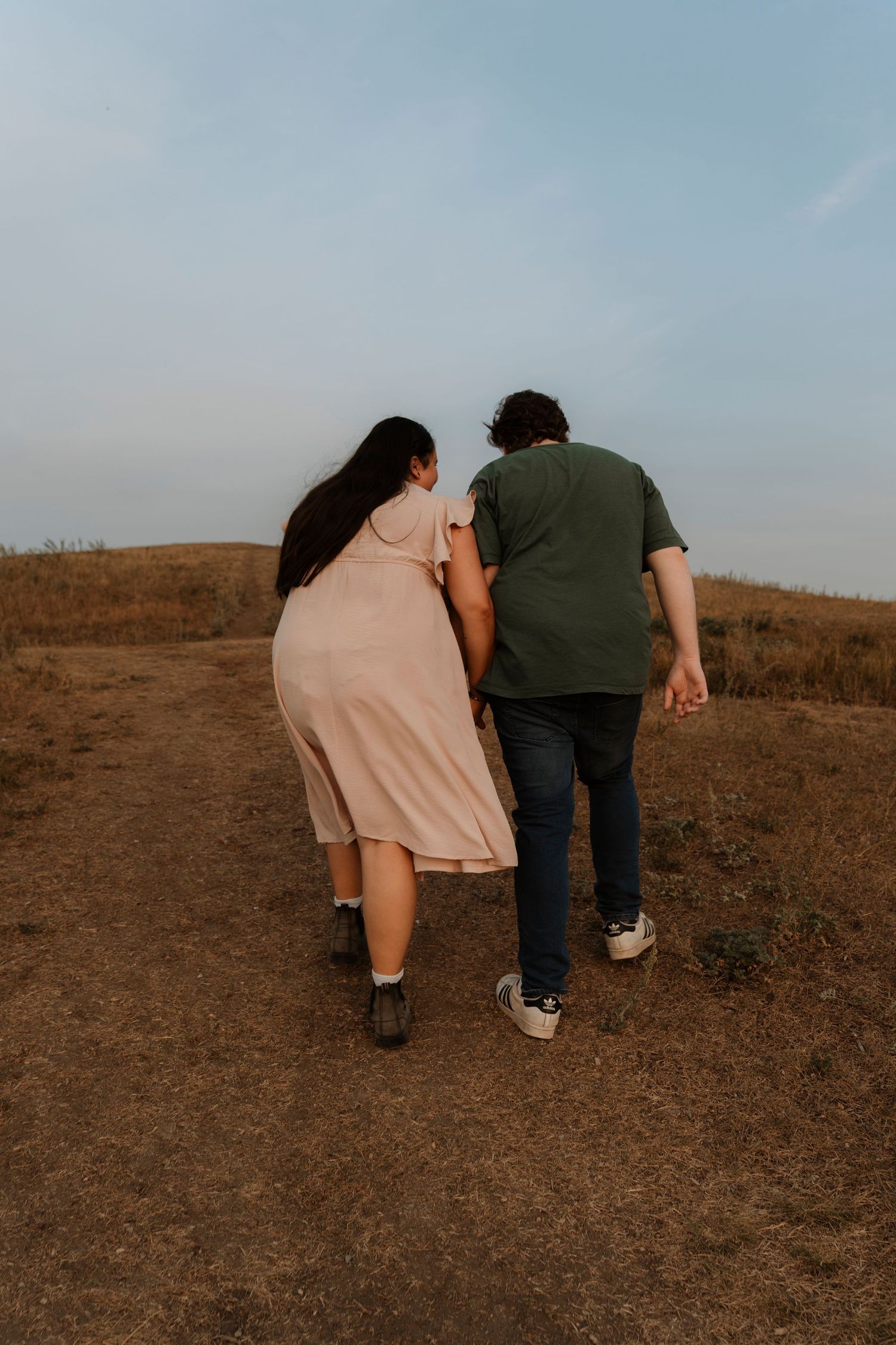 A couple walks hand-in-hand along a dirt path on a grassy hill under a pale blue sky.