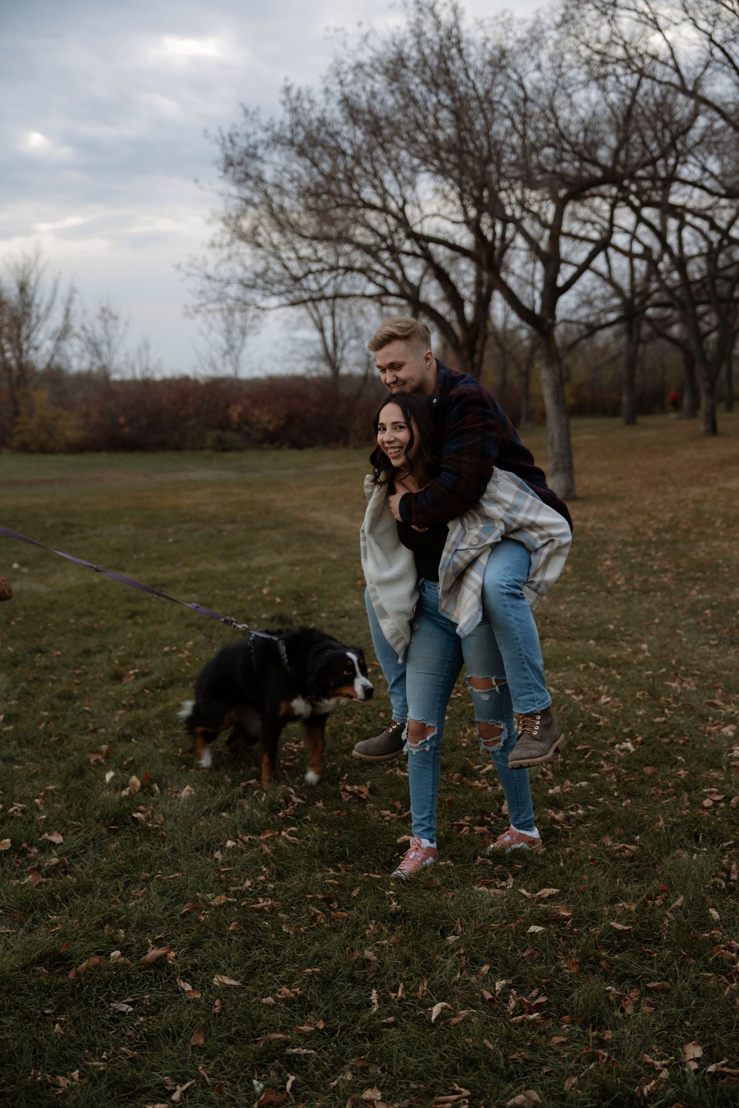Happy couple playing piggyback with their Bernese Mountain Dog in an autumn park.