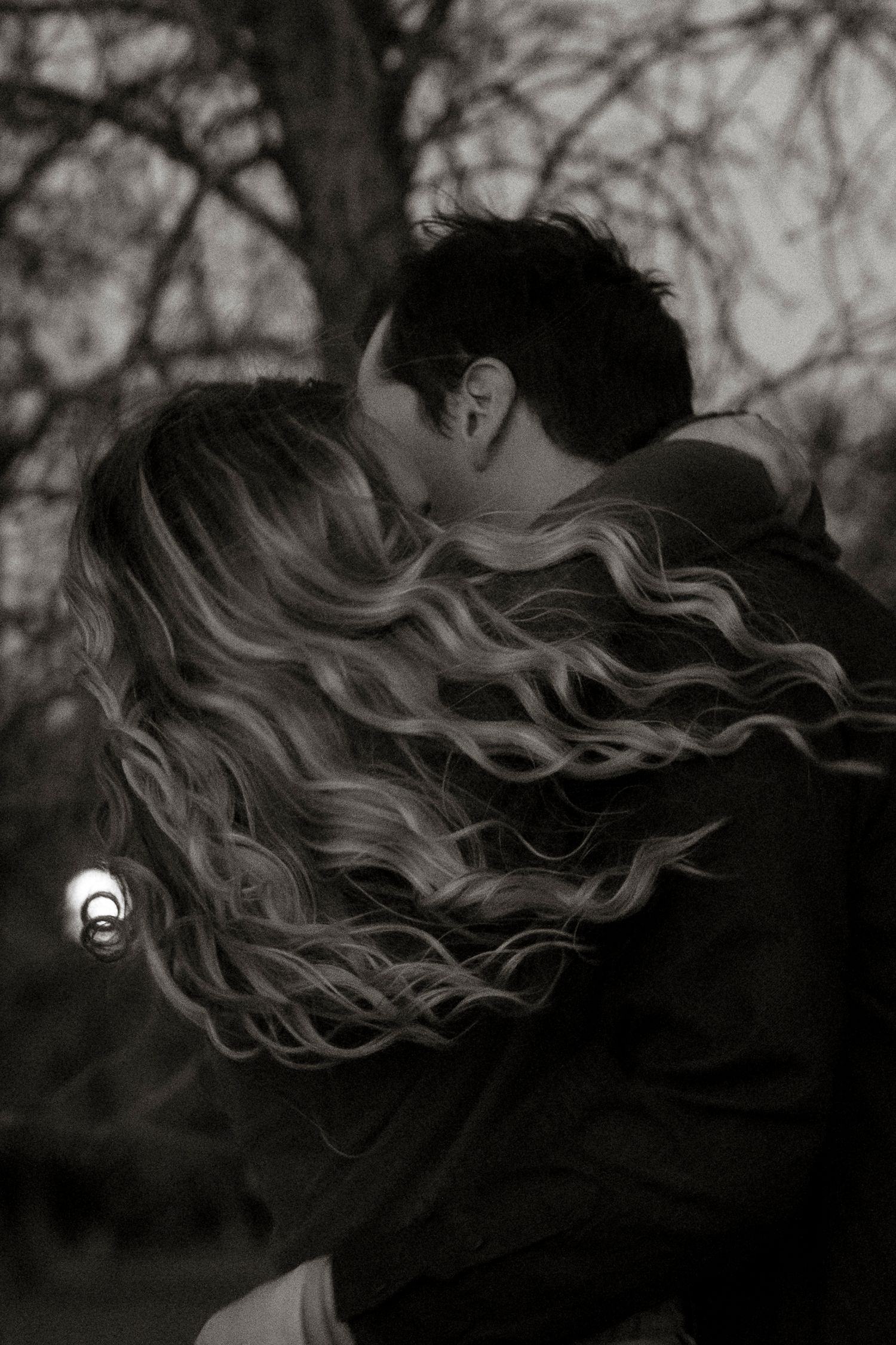 Black and white photo of a couple sharing a romantic kiss outdoors near a bare tree in winter.