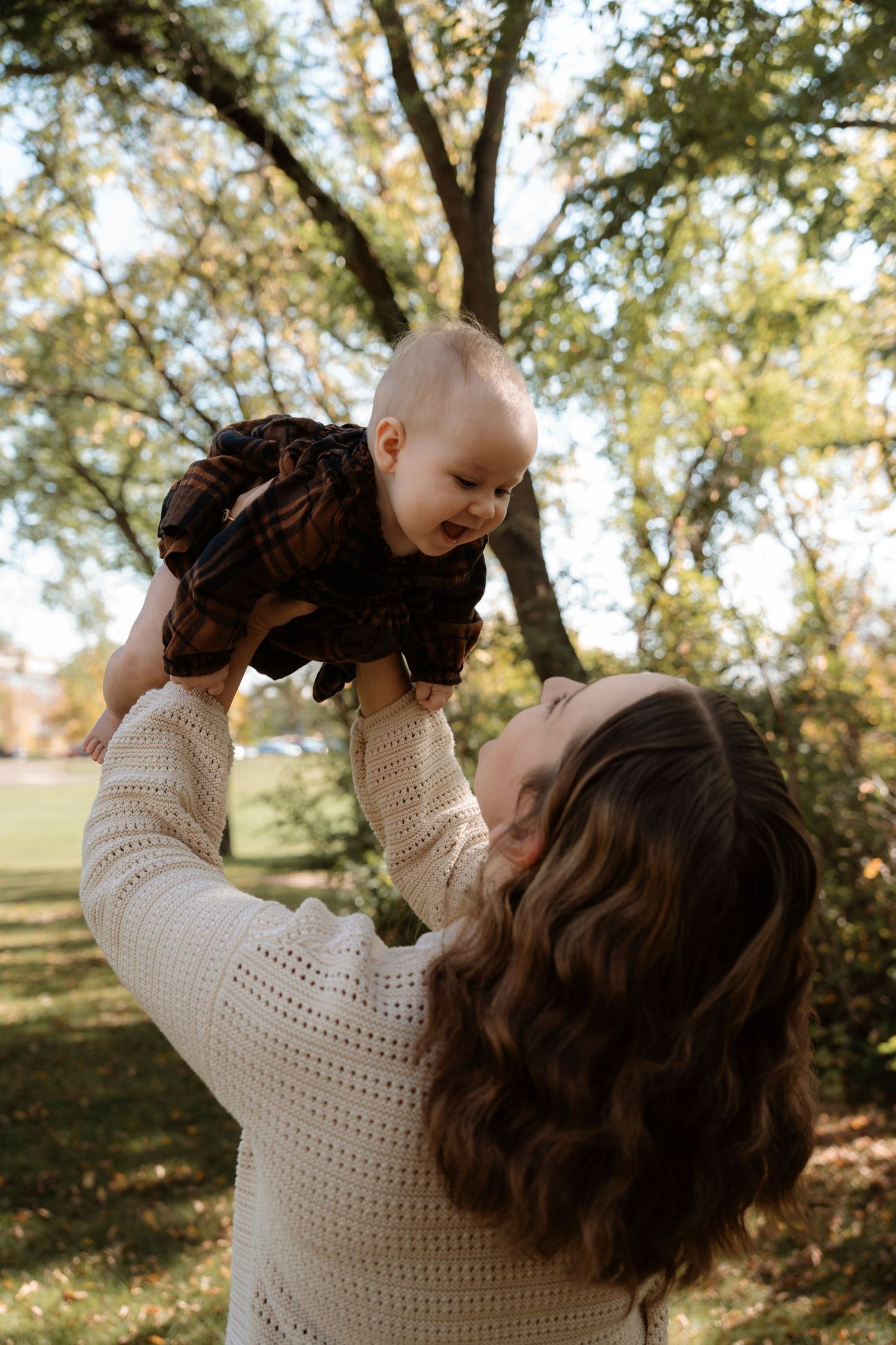 A mother lifts her smiling baby in a dark dress overhead outdoors, surrounded by lush green trees in warm sunlight.