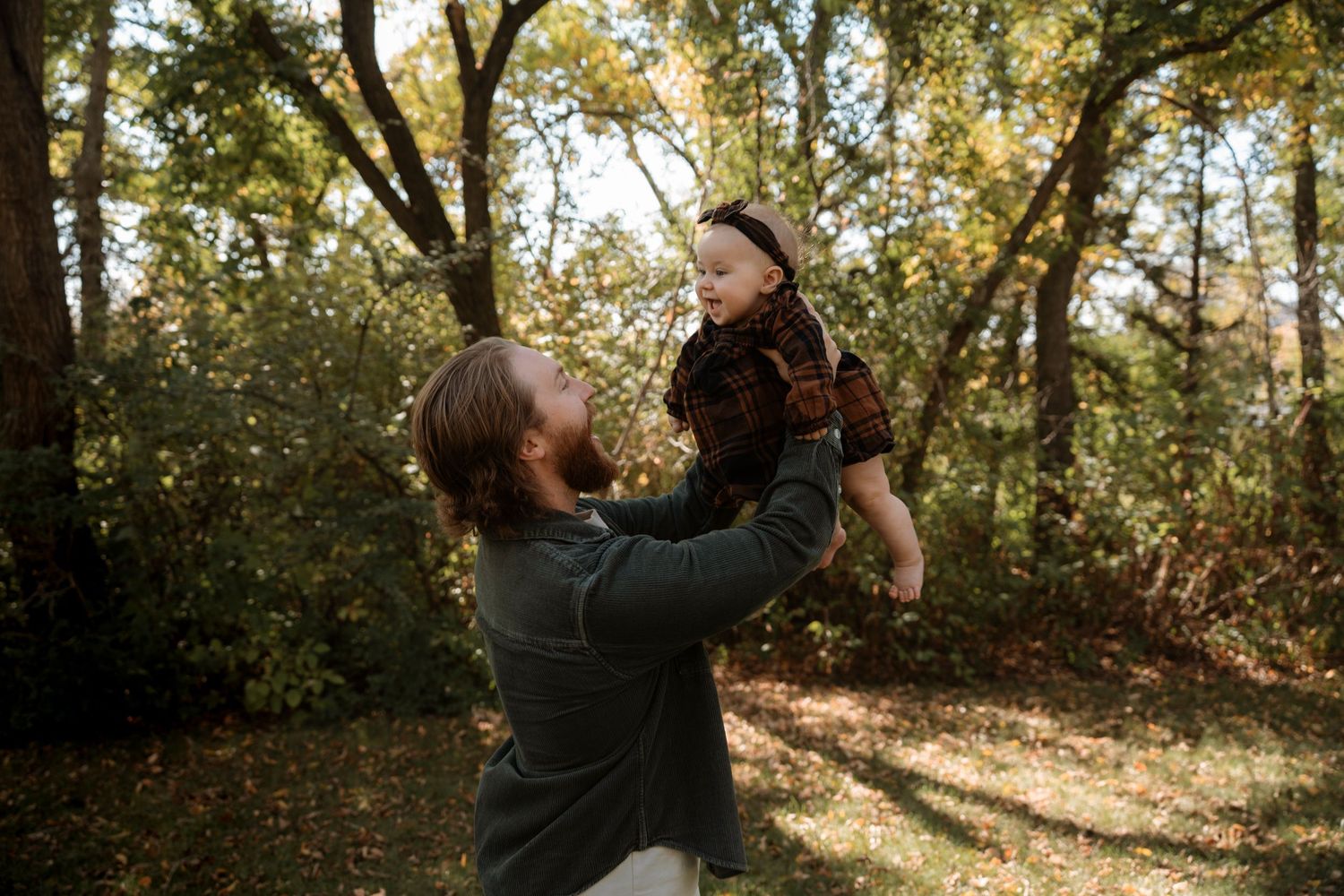 A bearded father joyfully lifts his smiling baby in a sunlit autumn forest setting.