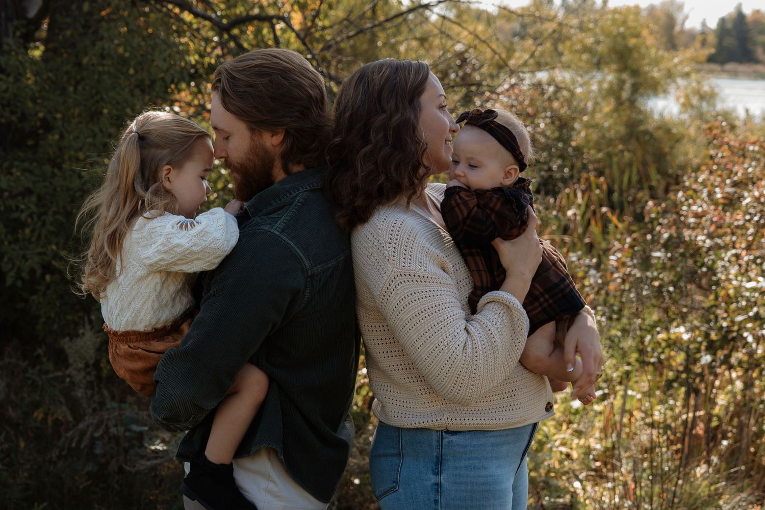 A couple holds two young children outdoors near a river surrounded by autumn foliage.