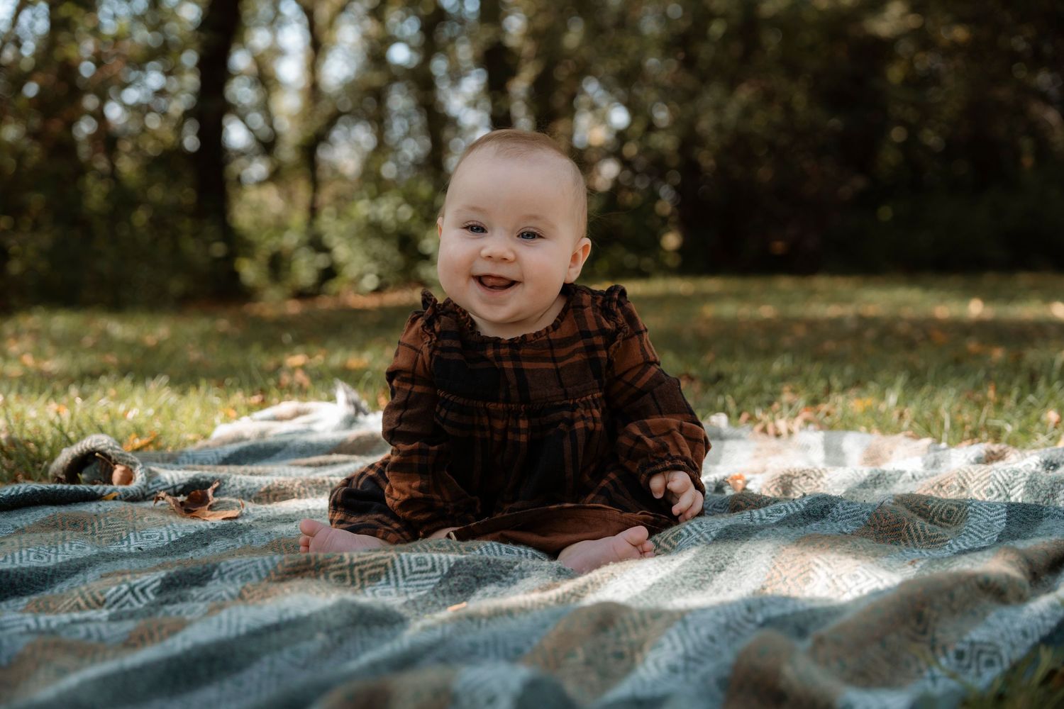 Smiling baby in brown plaid outfit sitting on a patterned blanket outdoors on green grass with trees in background.